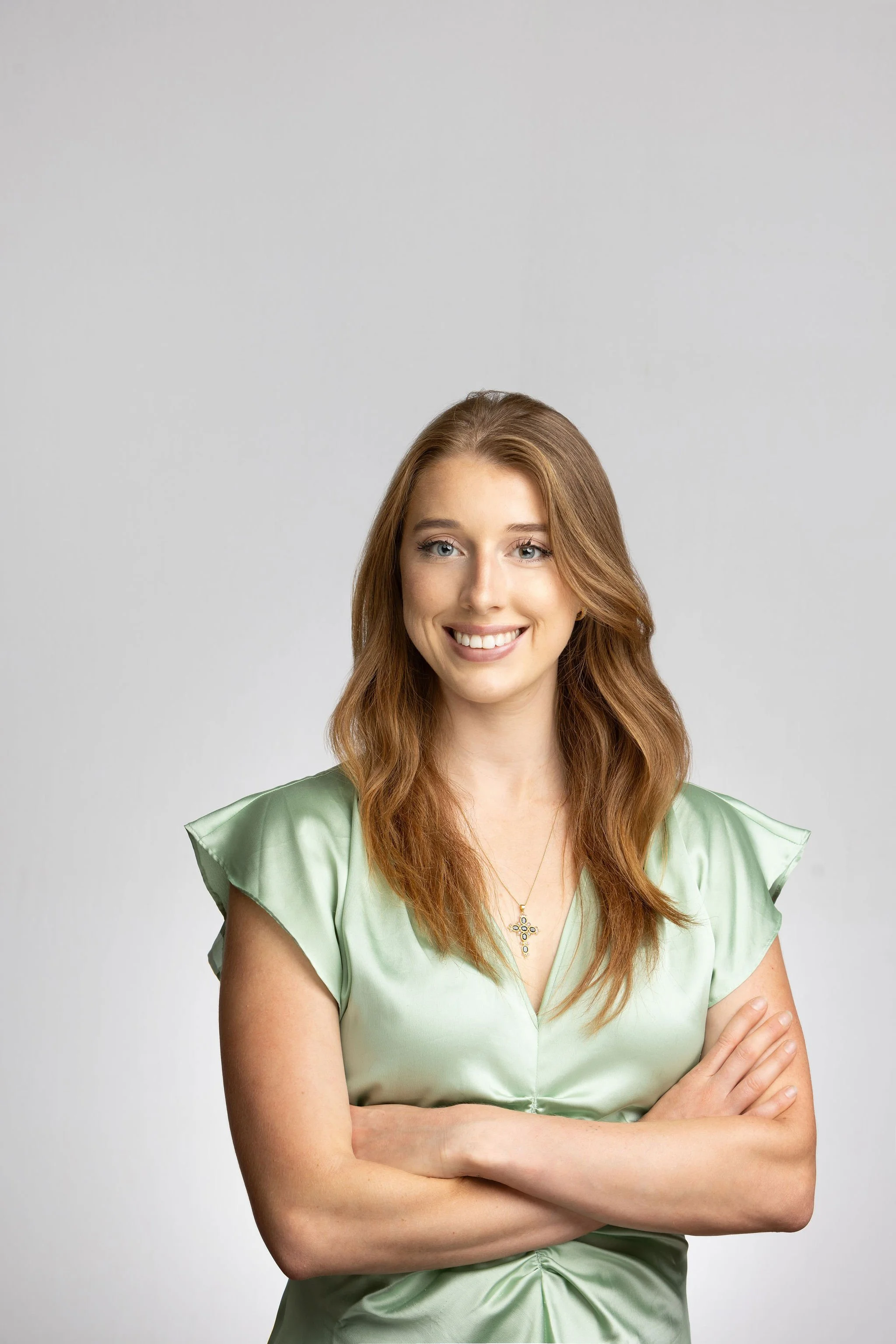 A young woman with long, wavy red hair smiling and crossing her arms, wearing a mint green satin dress with ruffled sleeves and a gold necklace with a decorative pendant, standing against a plain gray background.