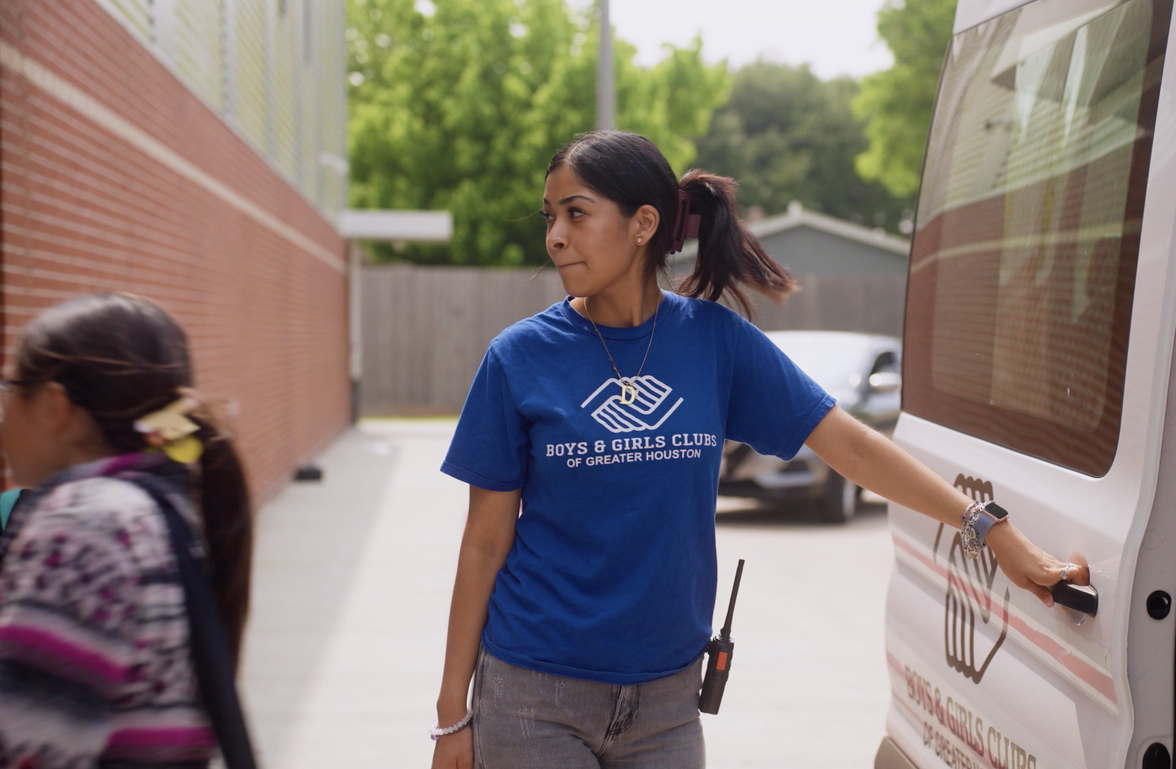 A young woman with dark hair tied back, wearing a blue Boys & Girls Clubs of Greater Houston t-shirt, standing outdoors near a vehicle with a sign. She is interacting with children, one with glasses and a colorful shirt, in a setting with green trees and a brick wall.