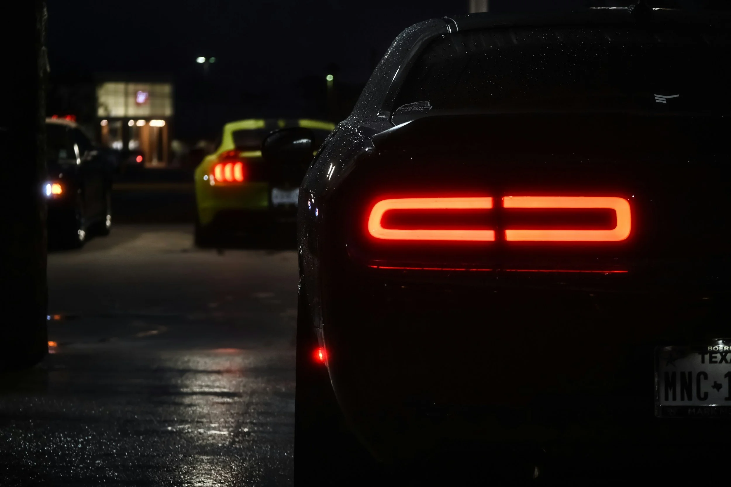Imagen de autos estacionados en la noche, destacando un coche negro con luces traseras encendidas, agua en la superficie y luces difusas en el fondo.