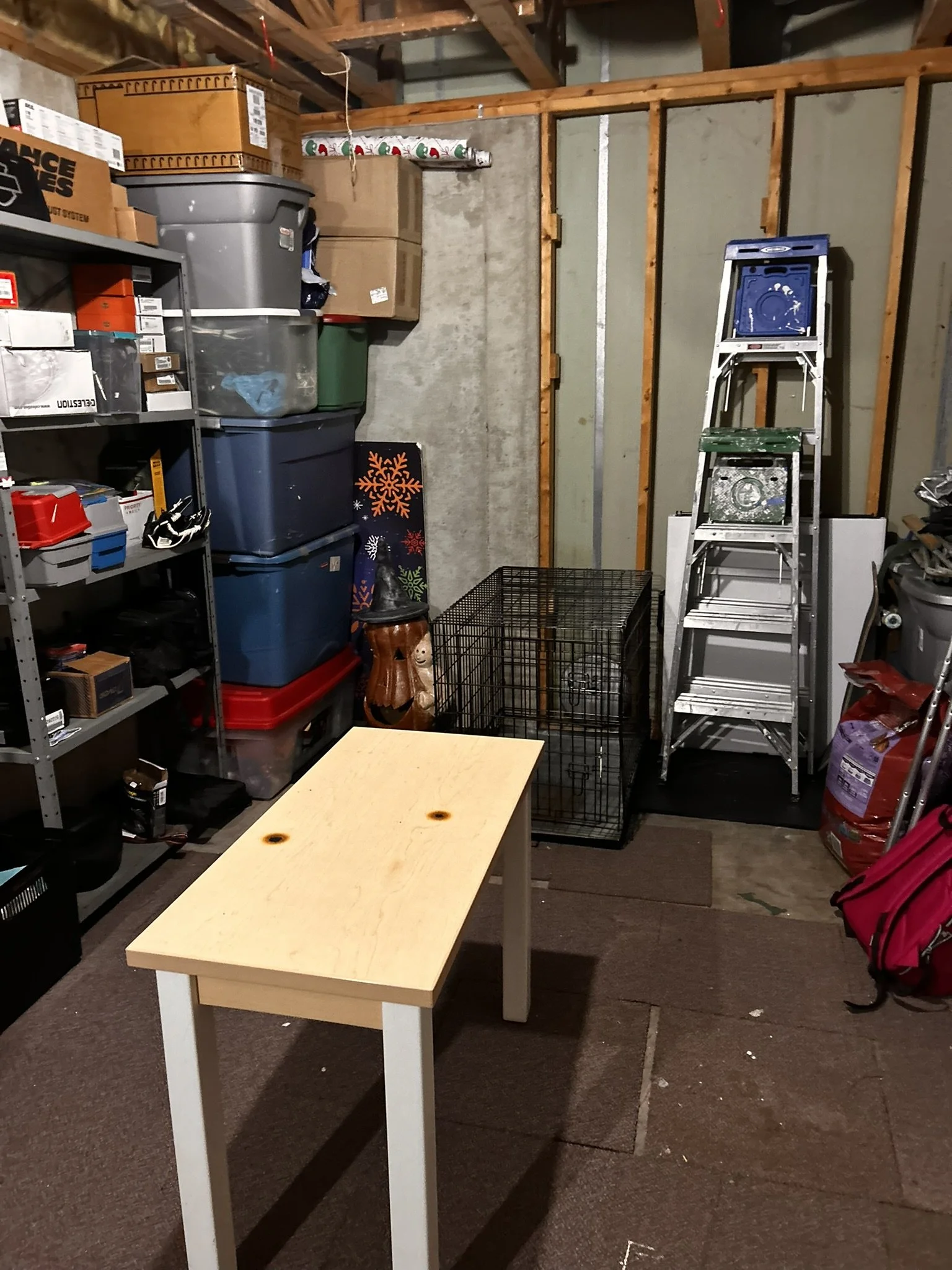 A cluttered garage with a beige worktable in the center, surrounded by shelves of storage bins, boxes, and miscellaneous items. A tall aluminum ladder and a black wire pet crate are also visible. The garage walls are unfinished with exposed wood framing and concrete.
