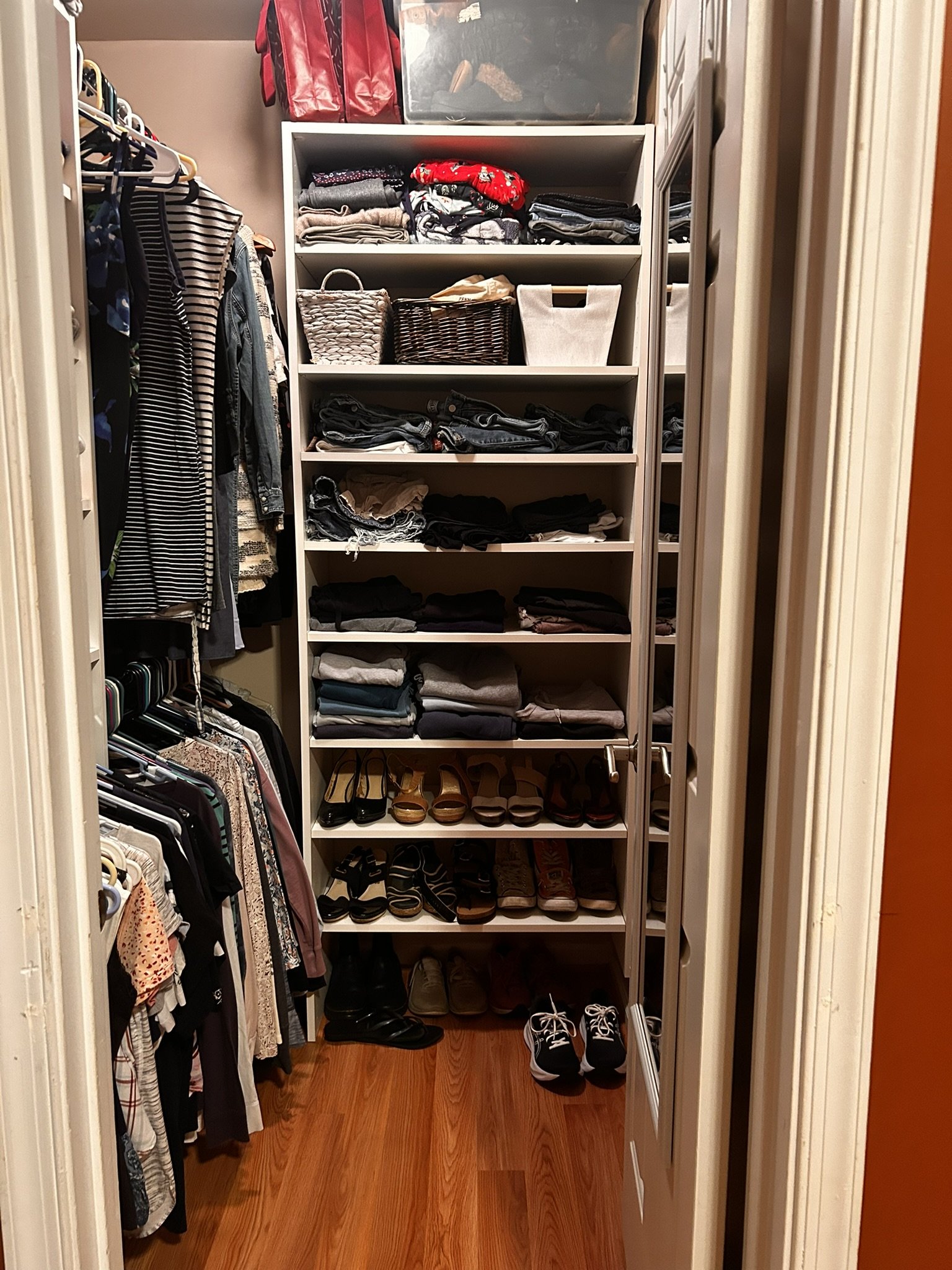A walk-in closet with white shelving units in the center filled with folded clothes, baskets, and boxes. On the left side, there are hanging clothes including shirts and dresses. Below the hanging clothes, there are more clothes and shoes. On the right side, there are additional shelves with shoes and folded clothes. The closet has a wood floor and is viewed from the doorway.