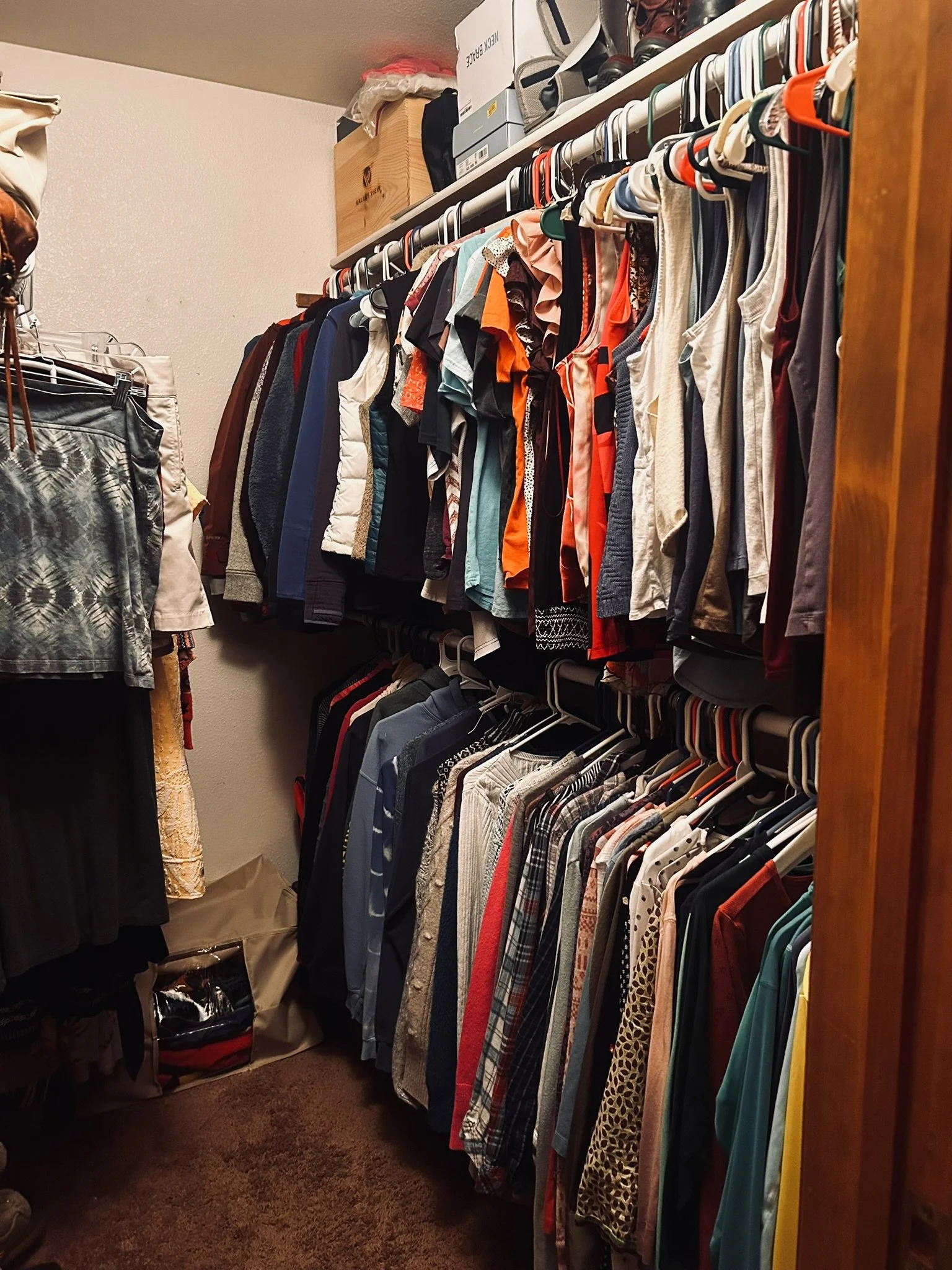 A walk-in closet with shelves and hanging clothes, including shirts, jackets, and dresses, along with some boxes and bags on the shelves.