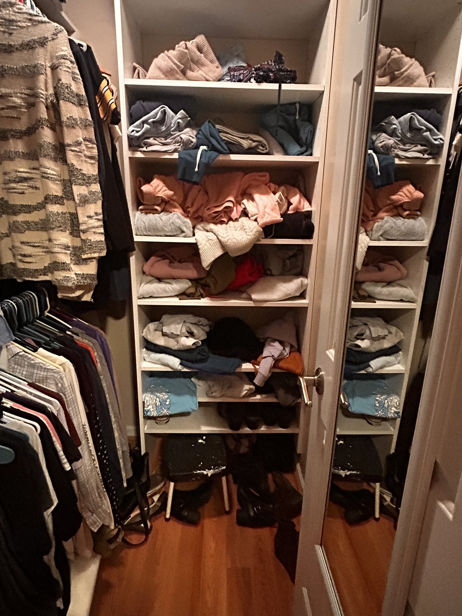 A closet with shelves filled with folded clothes, jackets, and accessories, with hanging shirts on the left side and a mirrored door on the right.