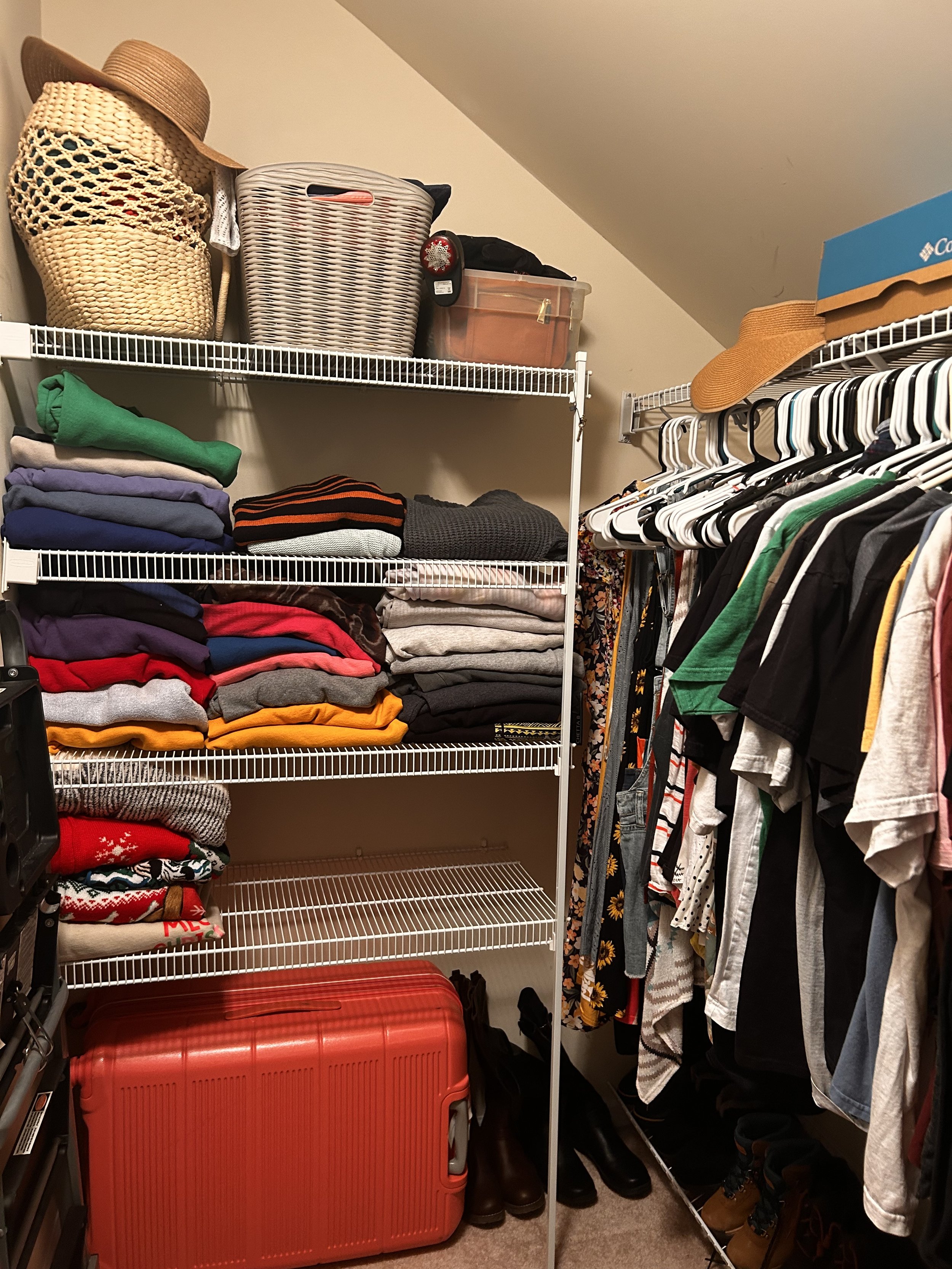 A closet with shelves filled with folded clothes, a large red suitcase, and hanging shirts and jackets on a rack.
