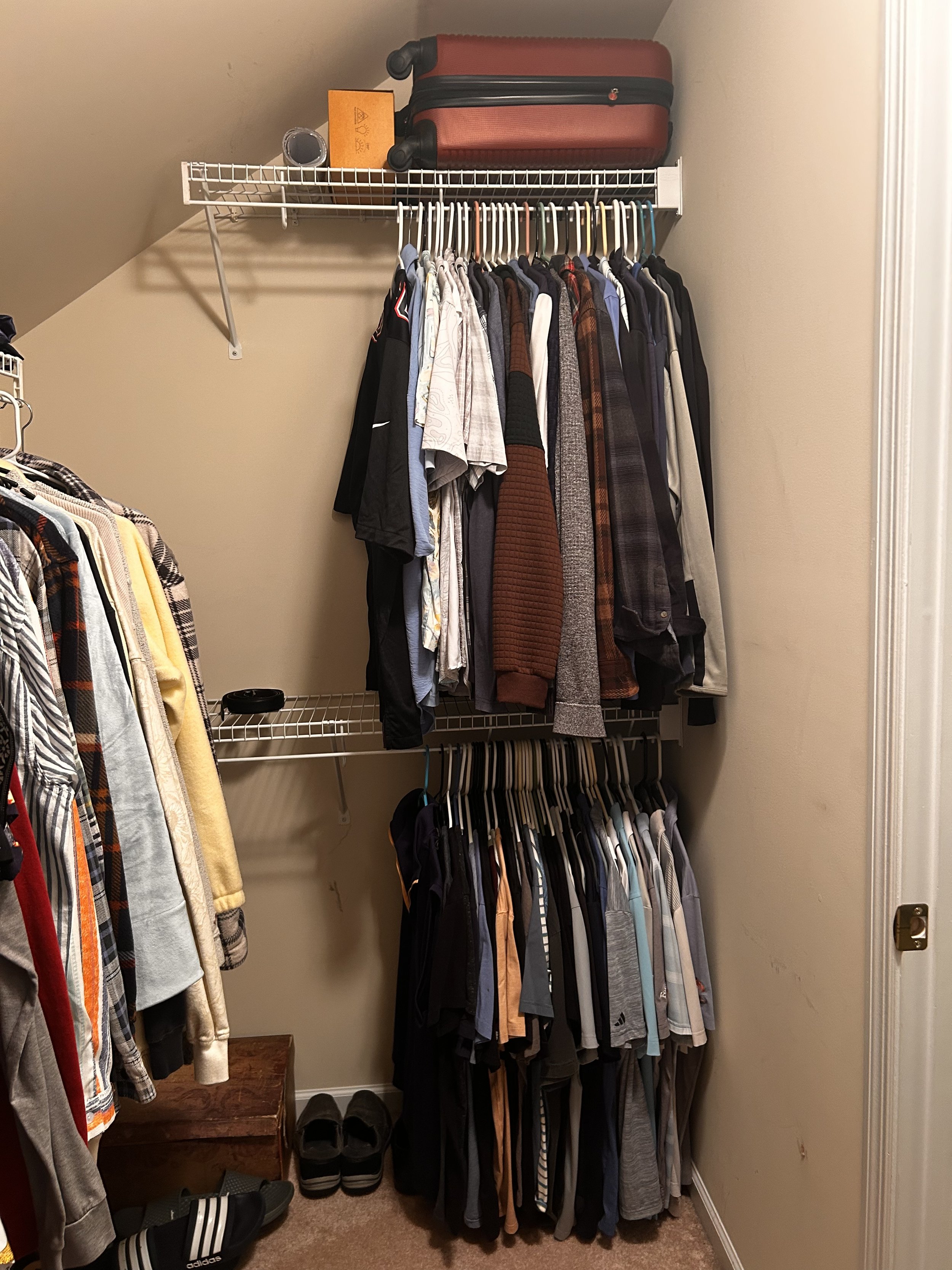 A closet with a metal wire shelf on the top holding a pink suitcase, a small orange box, and a black container. Below, a clothing rack filled with various shirts, jackets, and pants on hangers. To the left, part of another clothing rack with shirts and a yellow item. On the floor, shoes including black sneakers with white stripes and slippers, and a small wooden stool.
