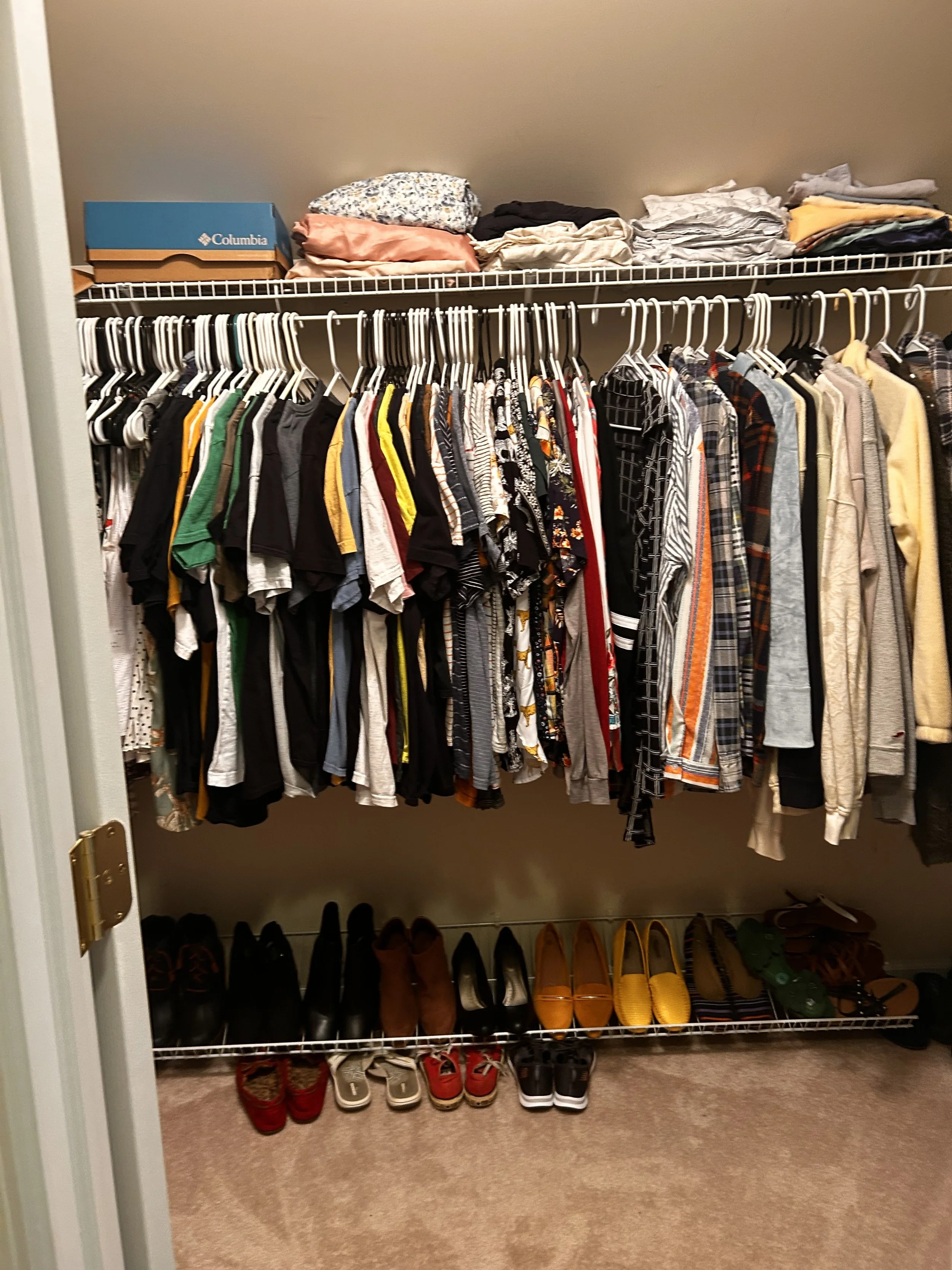 A closet with two white wire shelves. The top shelf contains folded clothes and a Columbia shoebox. The bottom shelf has hanging clothes including t-shirts, shirts, and jackets. Shoes are organized below the shelves, including boots, sneakers, and loafers.