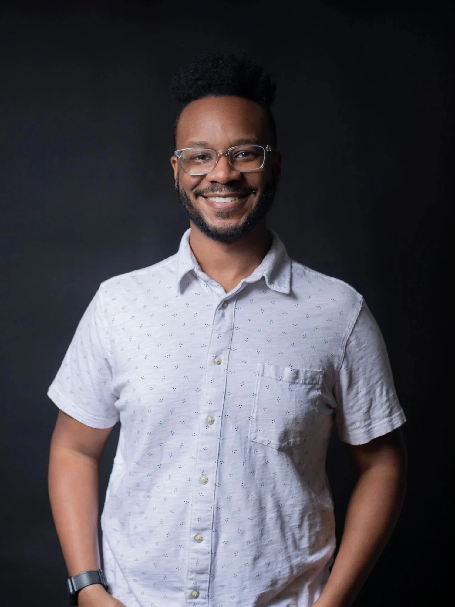 Smiling man with glasses and a beard in a white patterned shirt against a dark background.