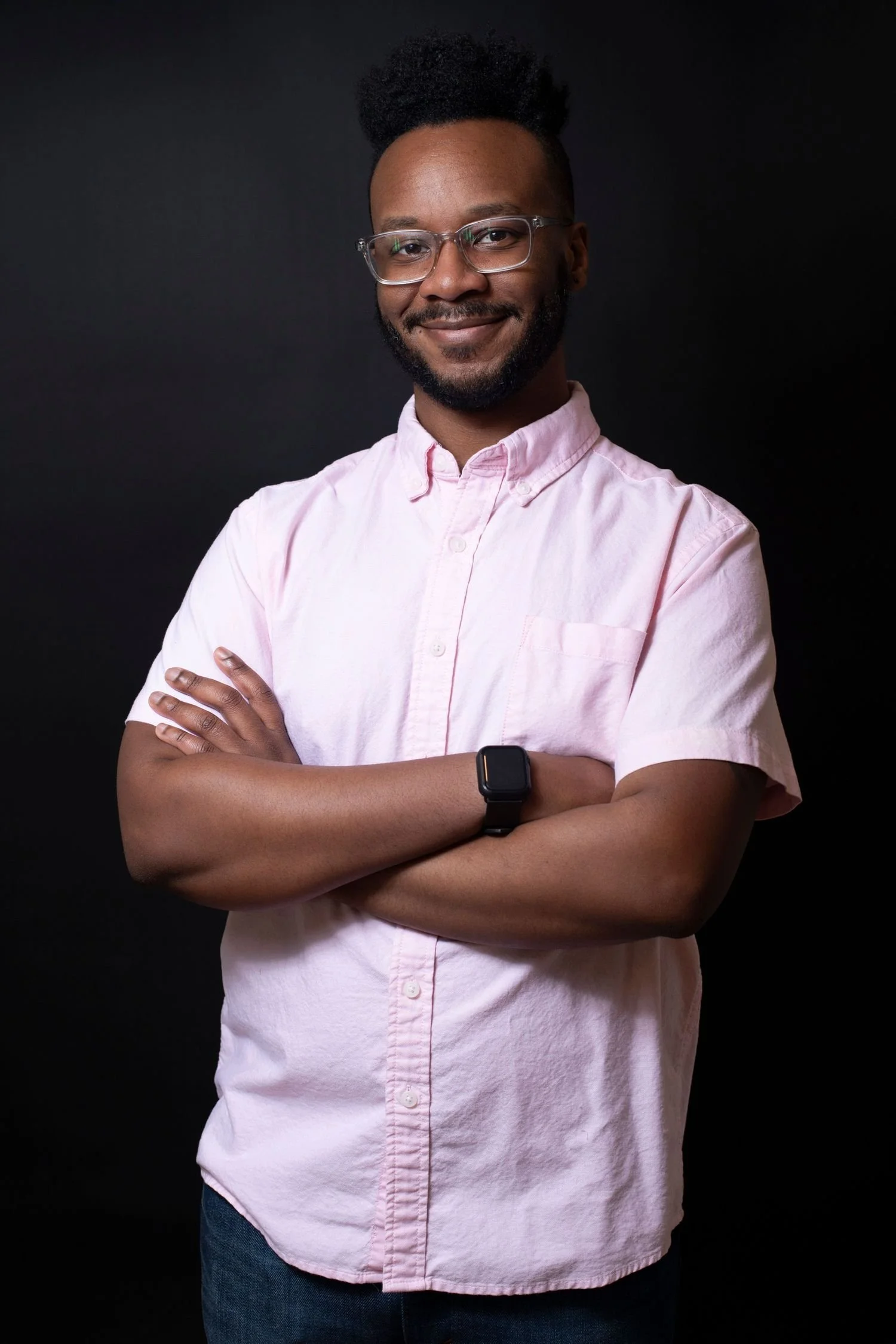 A young man with dark skin, glasses, a beard, and a fade hairstyle with natural hair on top, smiling and crossing his arms. He is wearing a light pink button-up shirt, a black smartwatch, and blue jeans. The background is black.