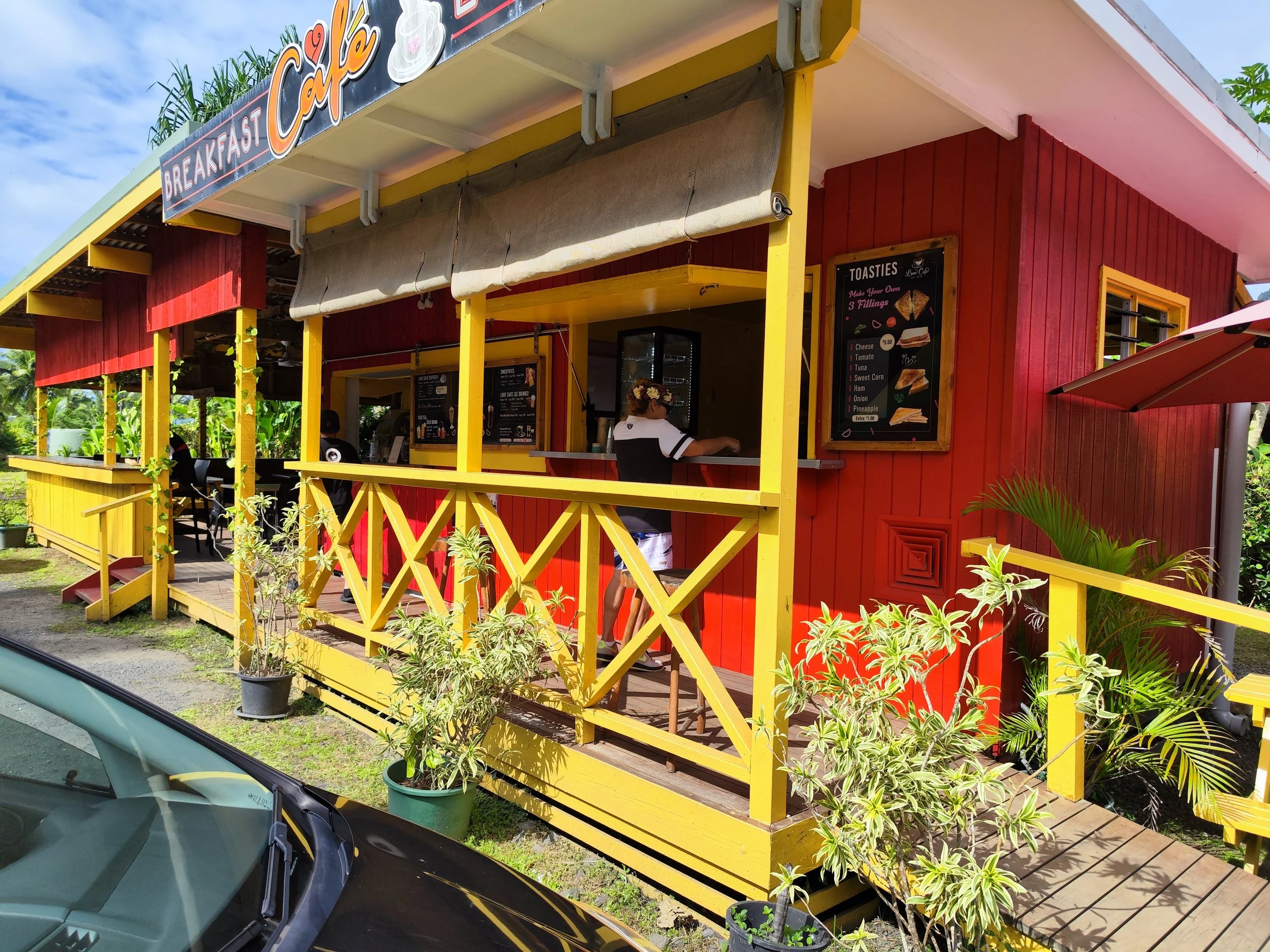 Colorful yellow and red cafe building with a sign offering breakfast and coffee. The outdoor seating area has potted plants and a menu board listing toast fillings. A woman stands inside behind the counter, with a person in front of the counter.