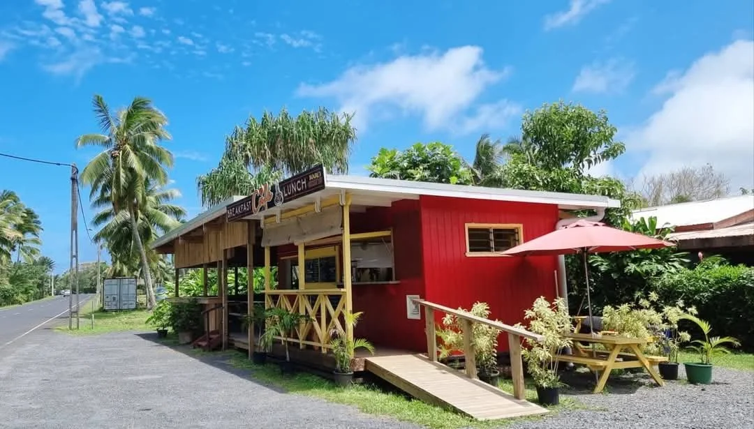 Small red wood cafe with yellow accents, outdoor seating with a pink umbrella, lush tropical trees, and a blue sky.
