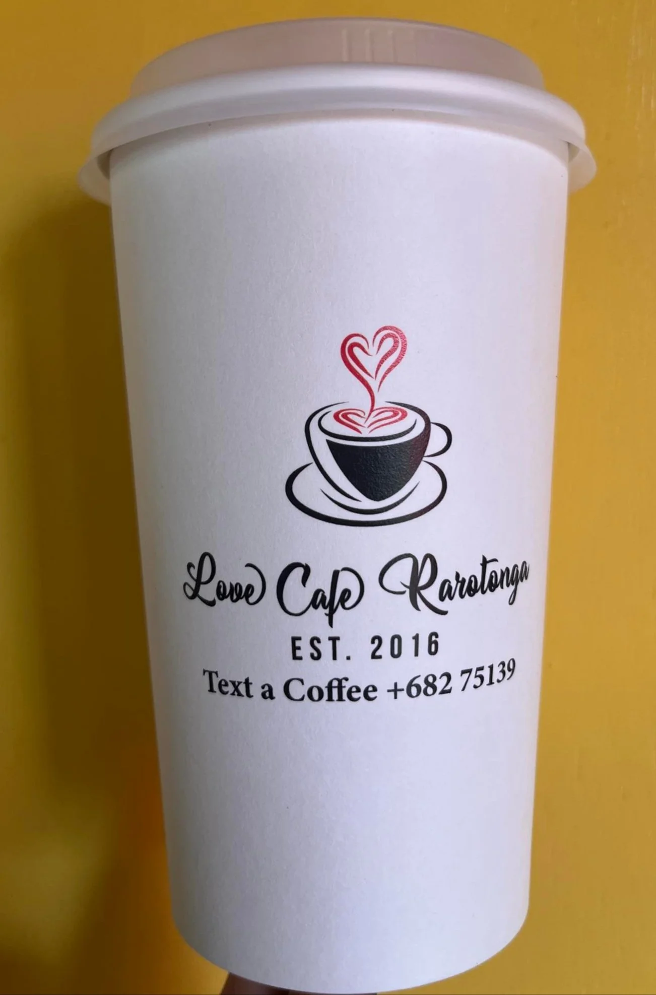 A white coffee cup sleeve with a logo of a steaming coffee cup in black and red, and the text 'Love Cafe Rarotonga EST. 2016'. Below, additional text reads 'Text a Coffee +682 75139'.