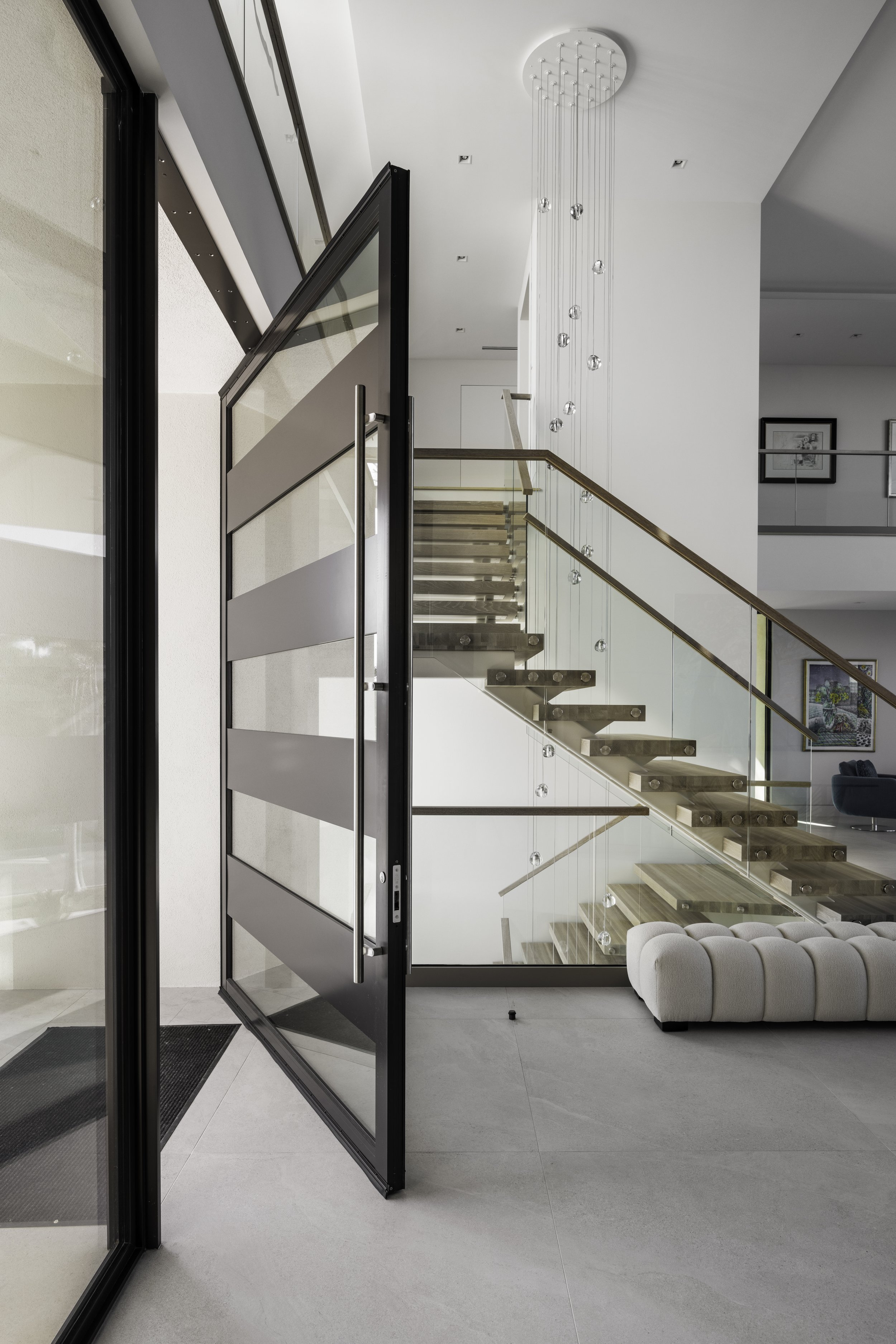 Modern interior with glass staircase, contemporary chandelier, and a beige sofa in a bright living space.