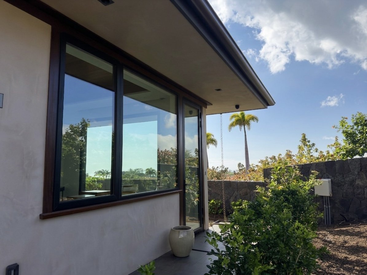 View of a house patio with a large window, a ceramic pot, green bushes, and tall palm trees in a sunny outdoor setting.