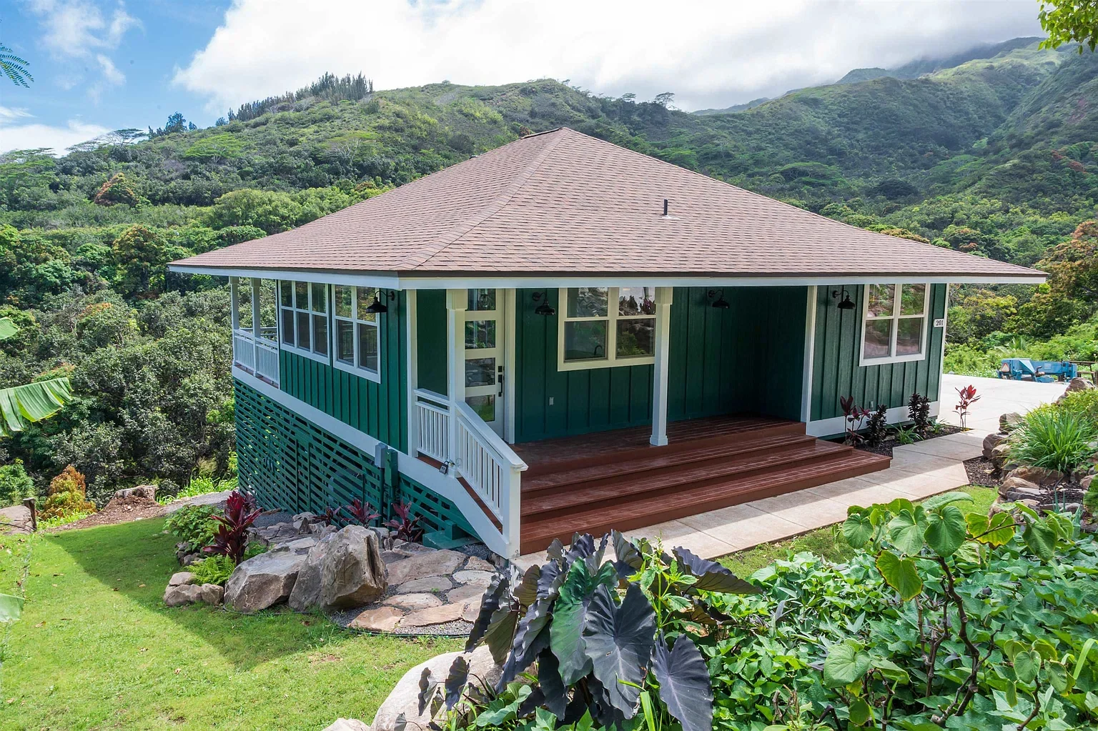 A green house with white trim and a brown roof is situated on a hillside with lush greenery and mountains in the background. It has a front porch with wooden steps and a garden with rocks and plants.