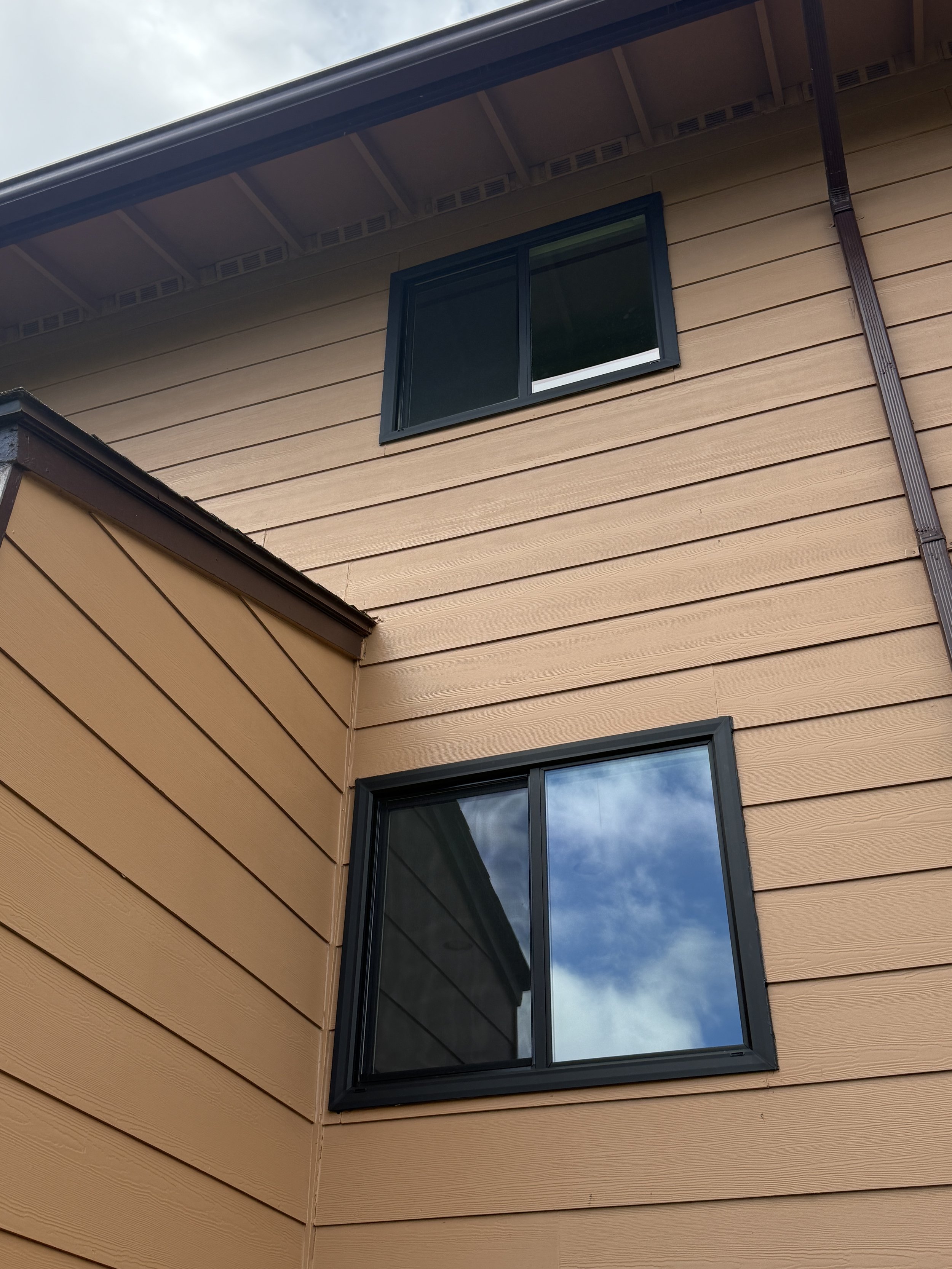 A modern house exterior with horizontal beige siding, black framed windows, and a brown gutter pipe on the corner.