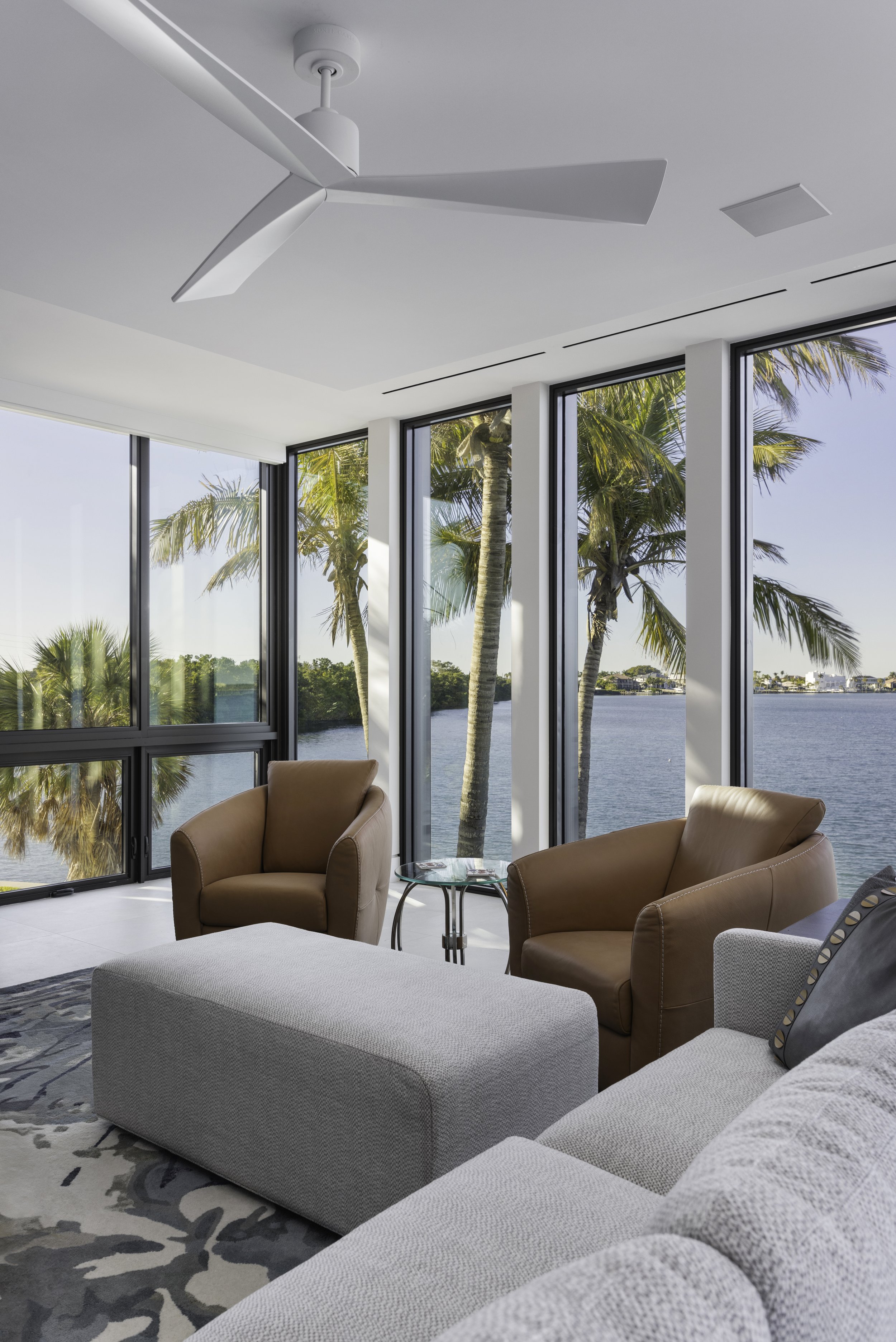 Living room with floor-to-ceiling windows overlooking a water view with palm trees, featuring brown armchairs, a light gray ottoman, and a patterned area rug.