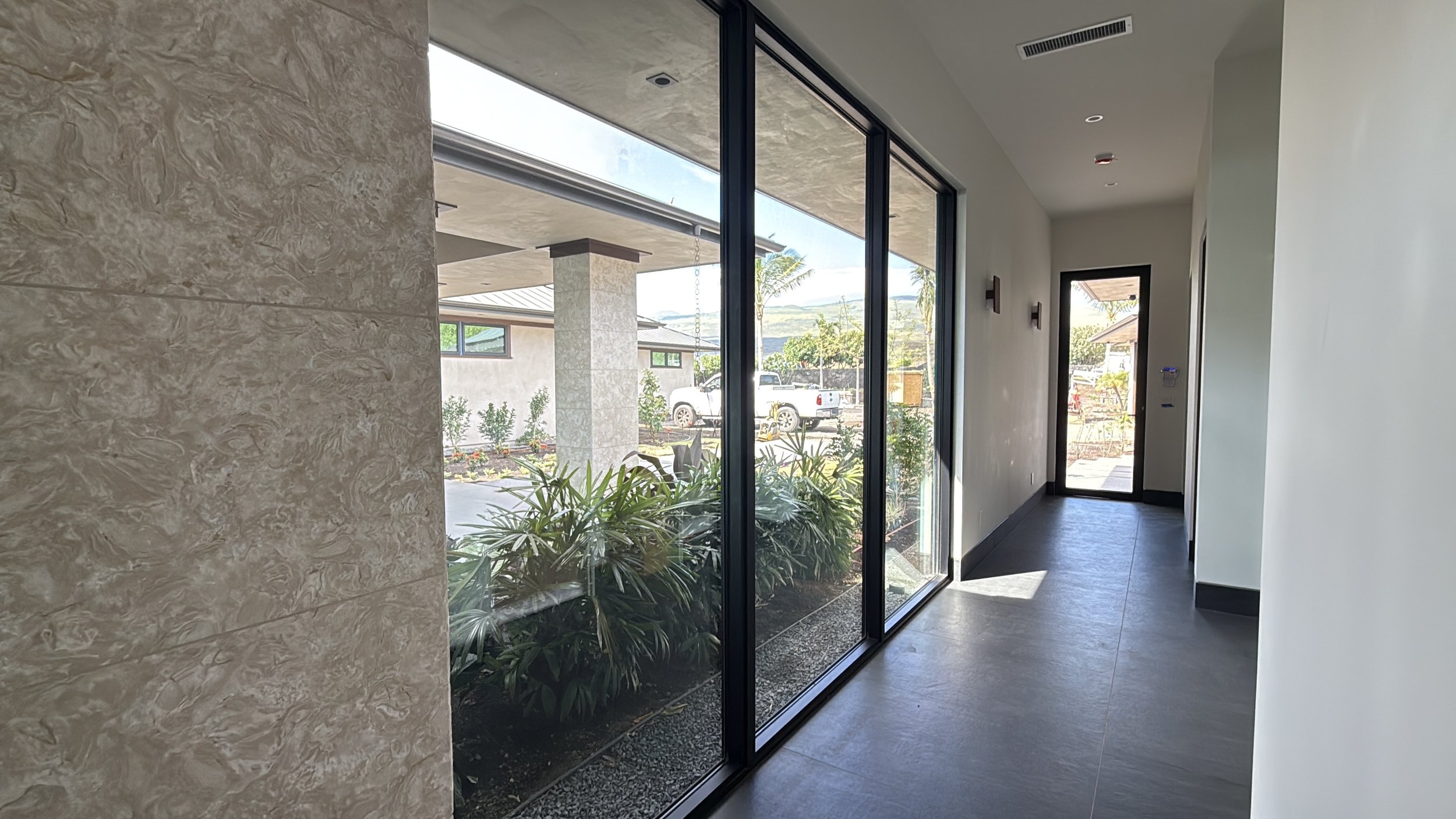 Interior hallway with large glass sliding door leading outside, view of a porch with plants, palms, and parked cars, modern design with textured wall and minimal decor.