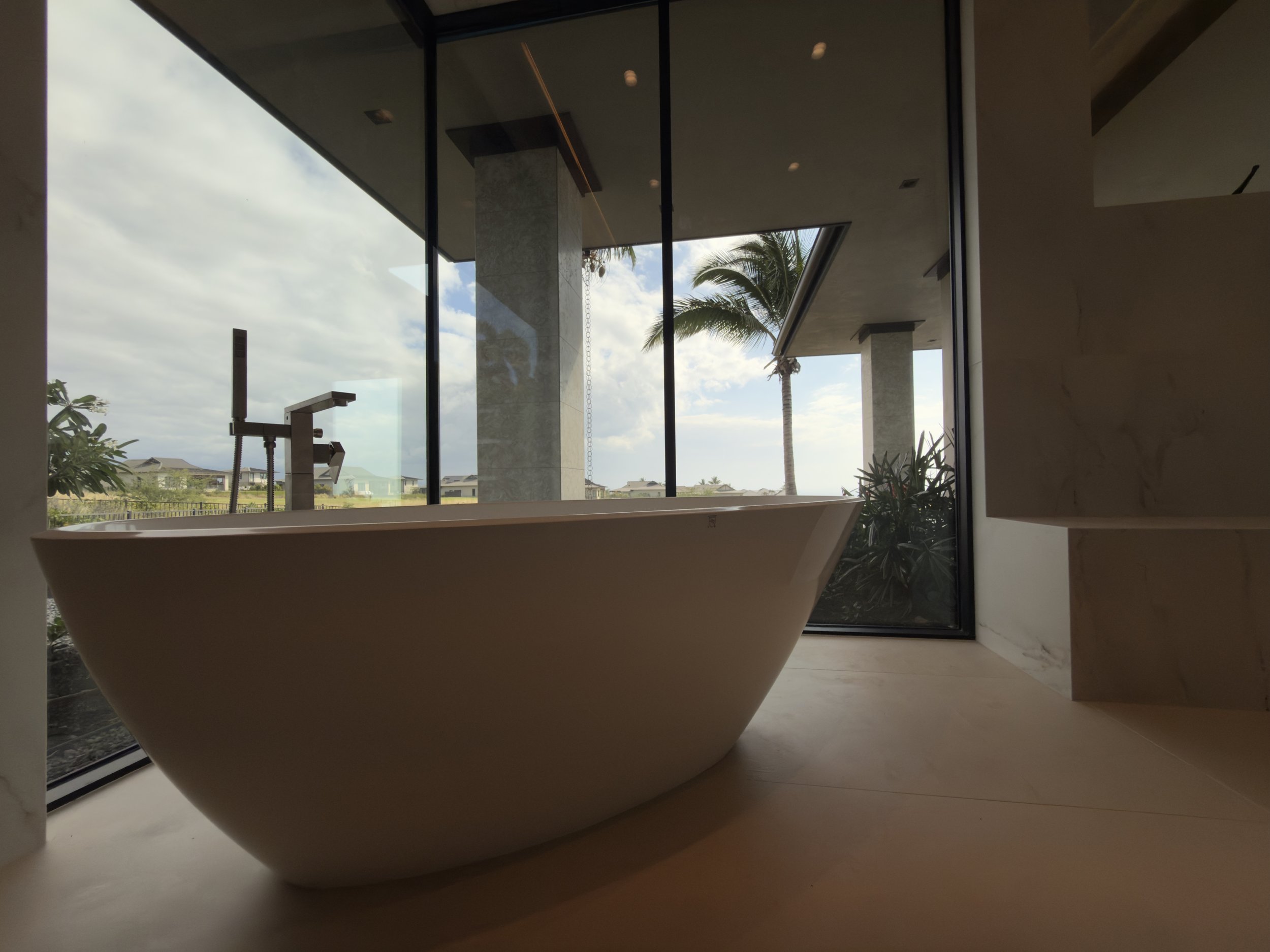 Modern bathroom interior with a freestanding bathtub near large floor-to-ceiling windows showing a view of palm trees and cloudy sky.
