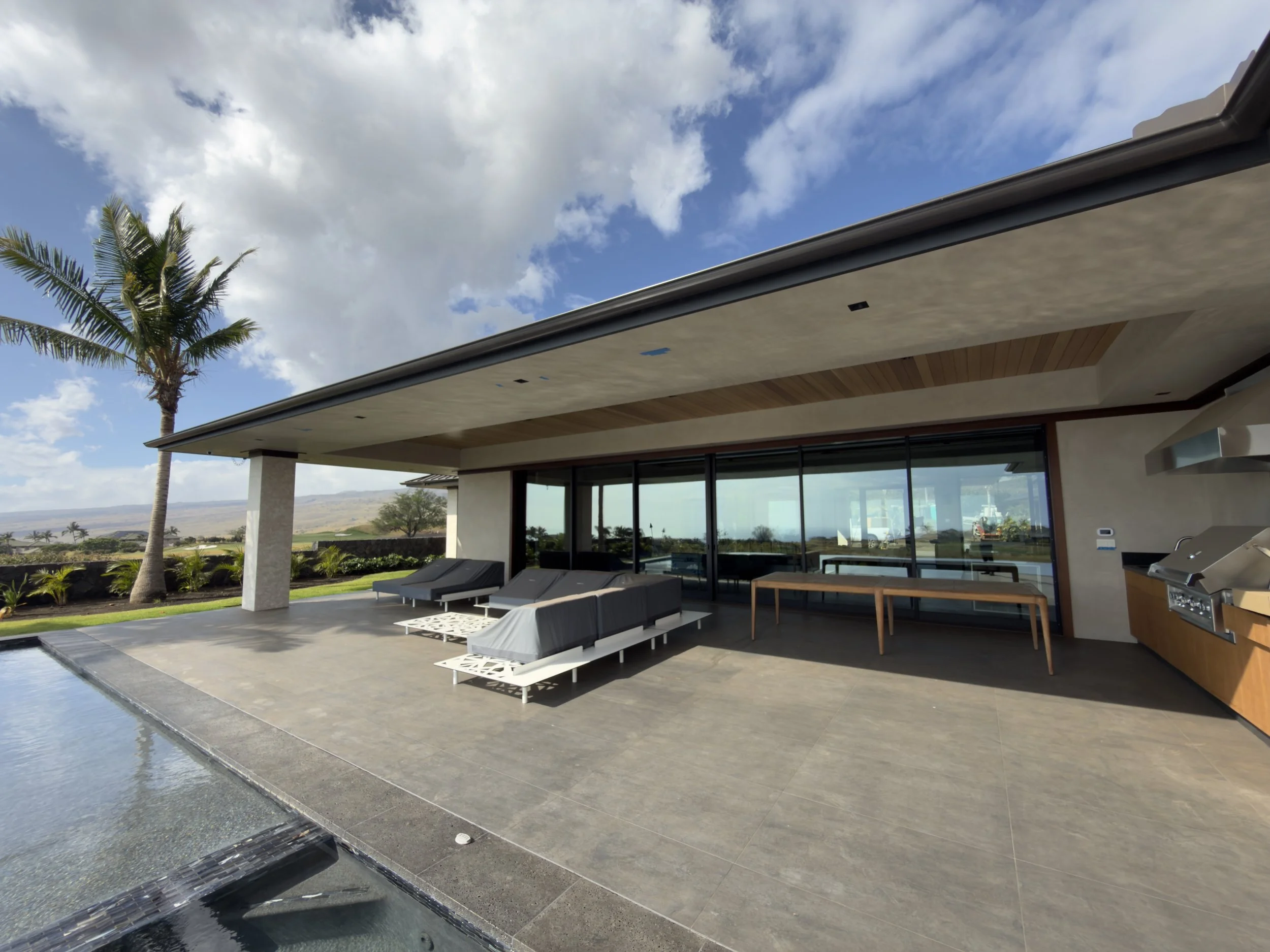 Modern house exterior with patio, pool, and outdoor lounge chairs, palm tree, and cloudy blue sky.