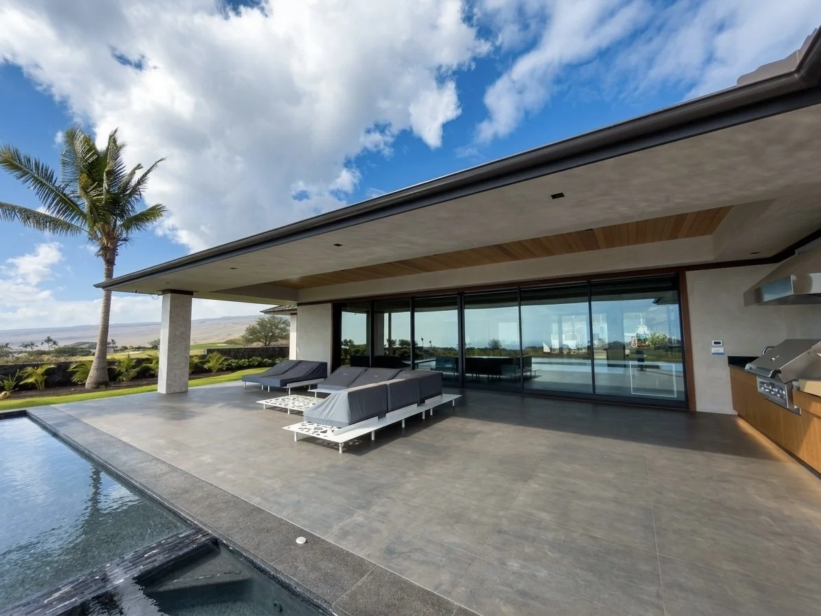 Modern outdoor patio with lounge chairs beside a pool, overlooking a landscape with palm trees and a blue sky with clouds.