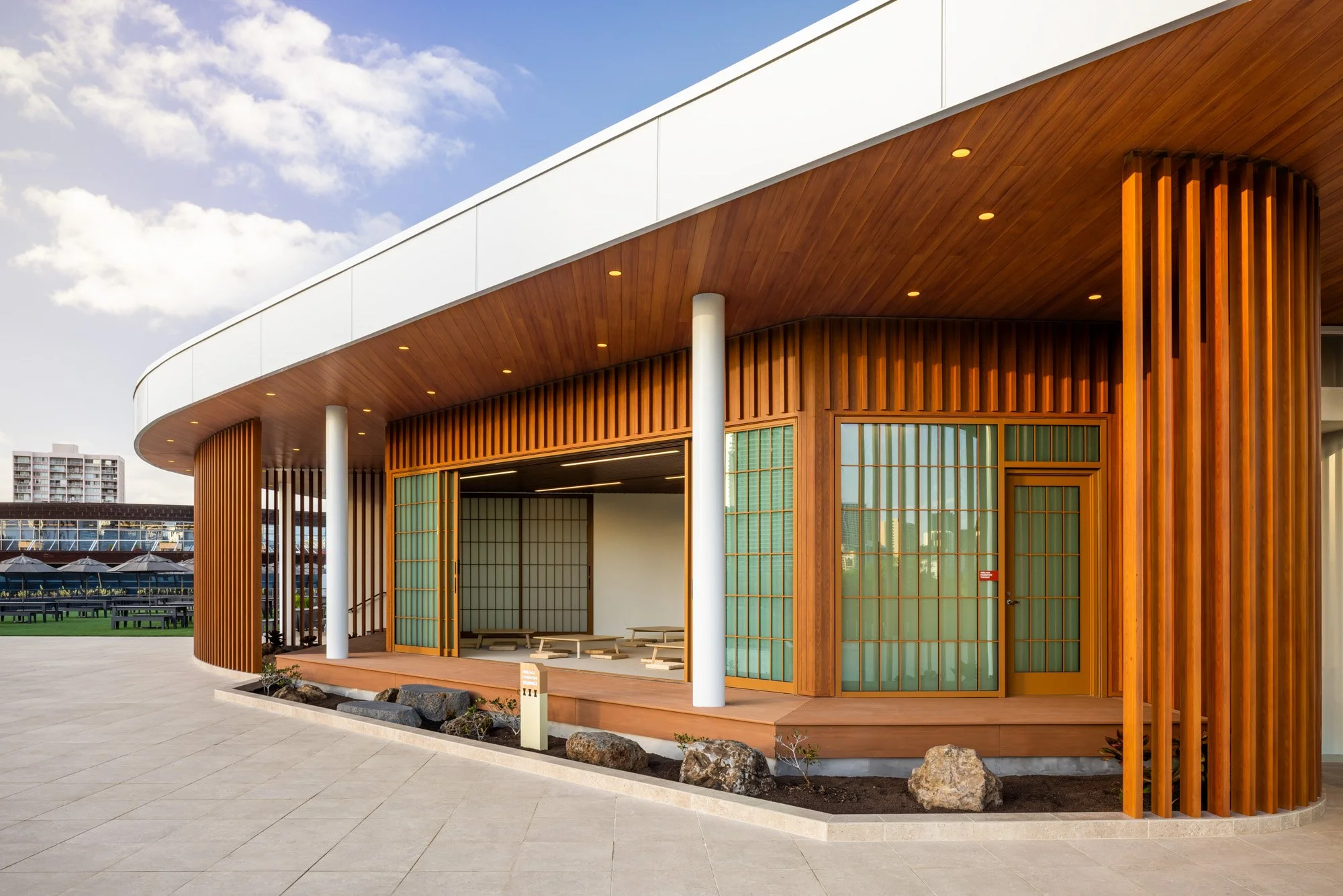 Modern building with a curved wooden facade, clerestory windows, and outdoor seating area, with a clear blue sky showing some clouds.