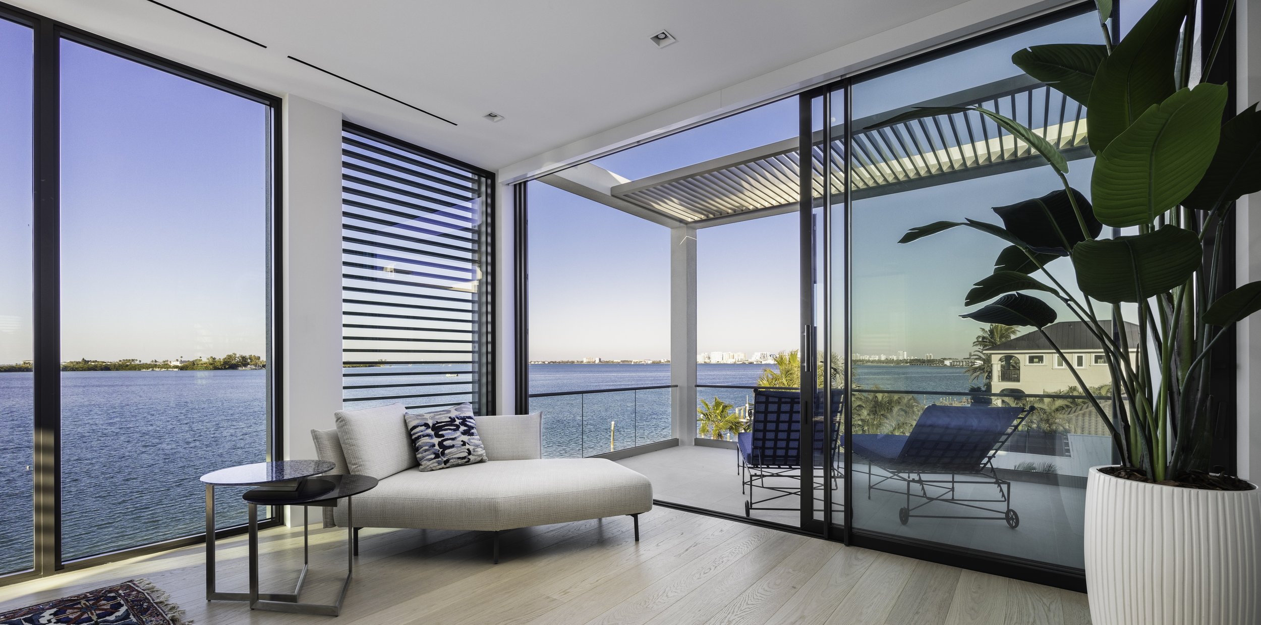 Interior of a modern apartment with large glass windows overlooking a body of water, with outdoor furniture and a potted plant inside.