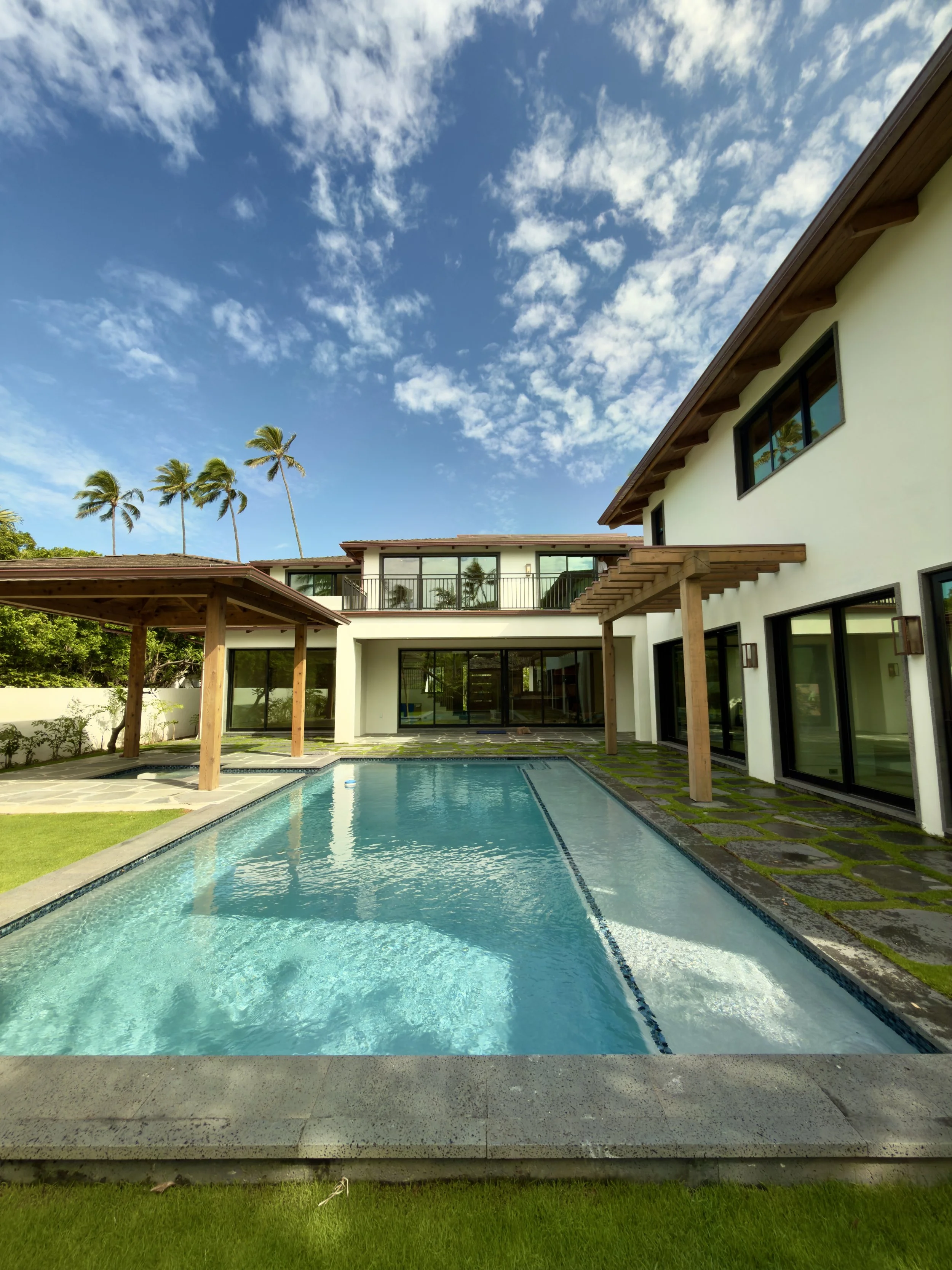 Modern house with backyard swimming pool, covered patio, and large glass windows under a blue sky with scattered clouds.