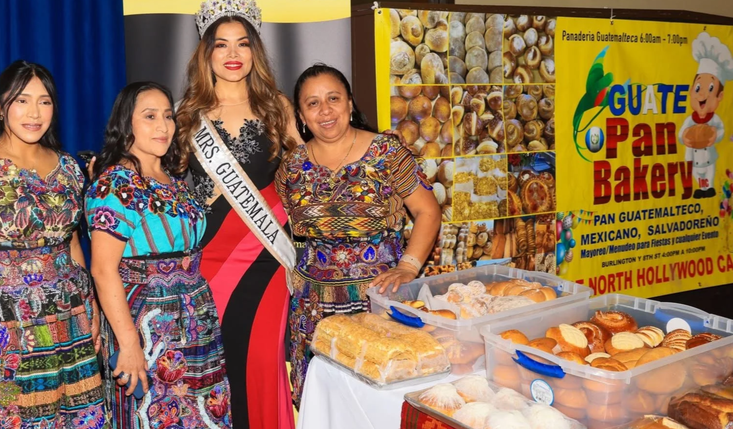 Four women standing behind a table with baked goods at a Guatemalan bakery stand during an event. One woman is wearing a crown and a sash that reads 'Mrs. Guatemala.' The table displays various bread and pastries, with a bright yellow banner in the background promoting a Guatemalan bakery.