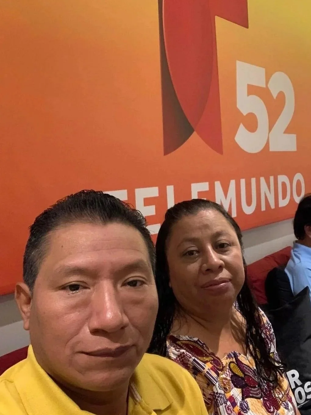 Two people taking a selfie in front of an orange wall with the logo of Televisa and the number 52 in white text.