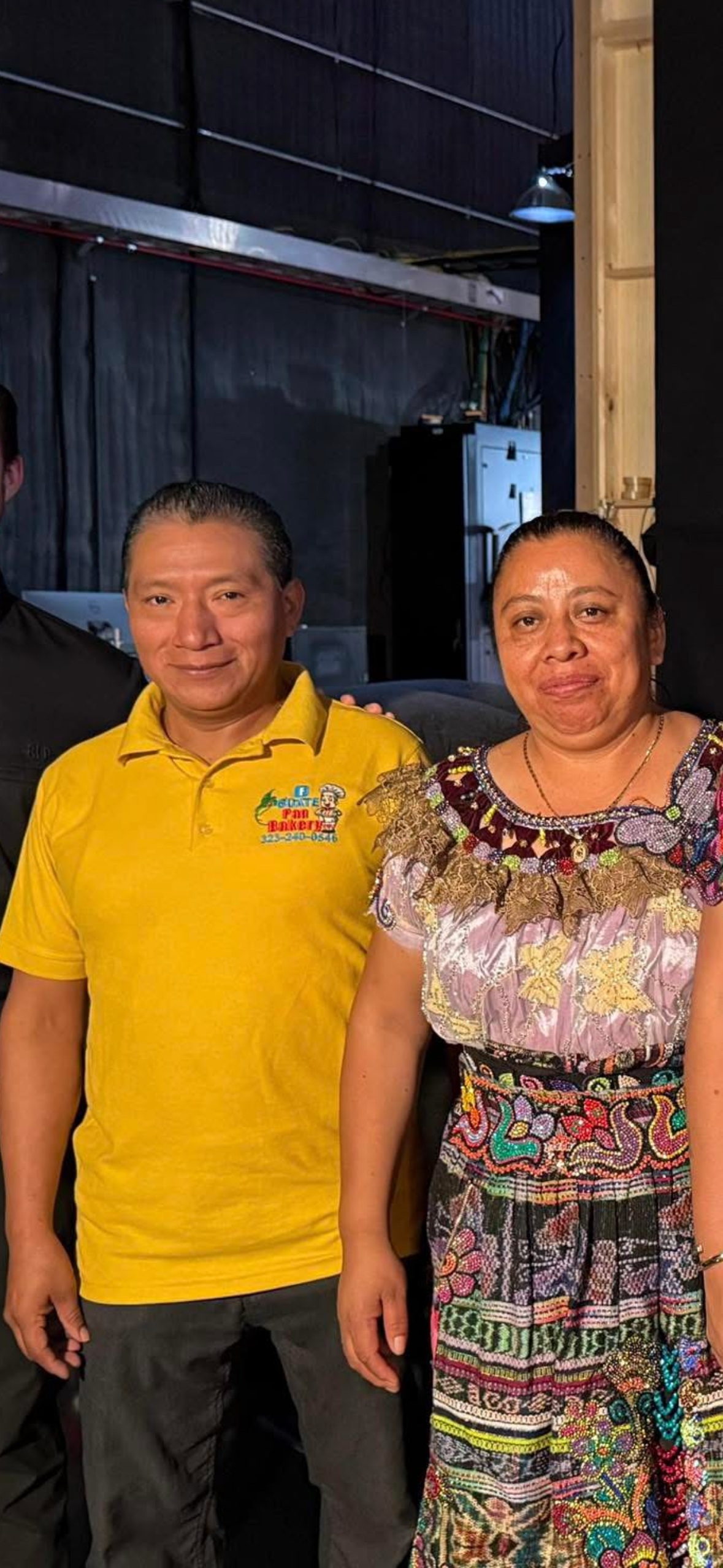 Two people standing indoors, posing for a photo. The person on the left is a man wearing a yellow polo shirt with a bakery logo and phone number. The person on the right is a woman dressed in colorful, traditional embroidered clothing.