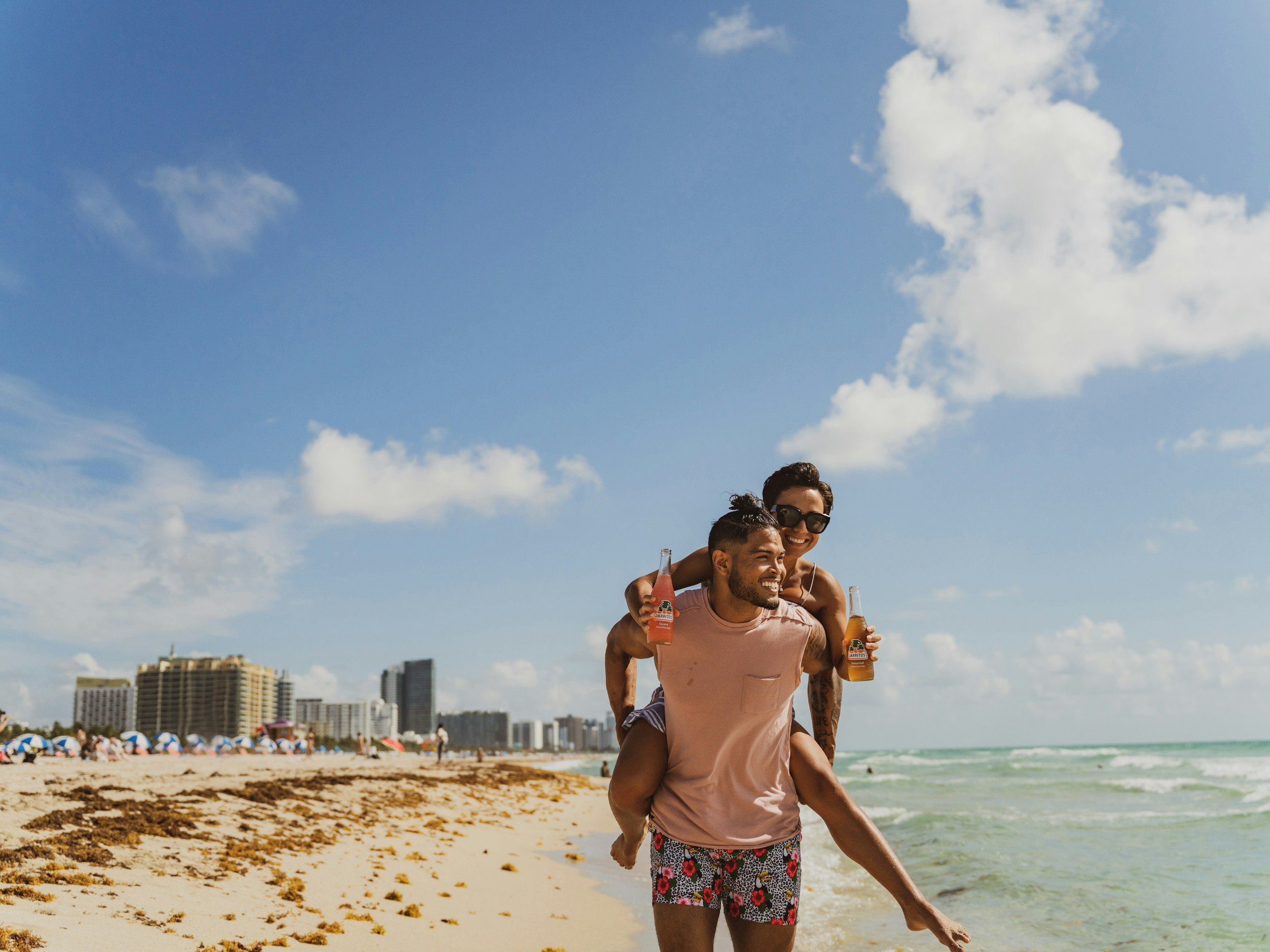 Two friends enjoying a day at the beach, with one giving the other a piggyback ride while holding drinks, against a backdrop of beach umbrellas, buildings, and a blue sky with clouds.