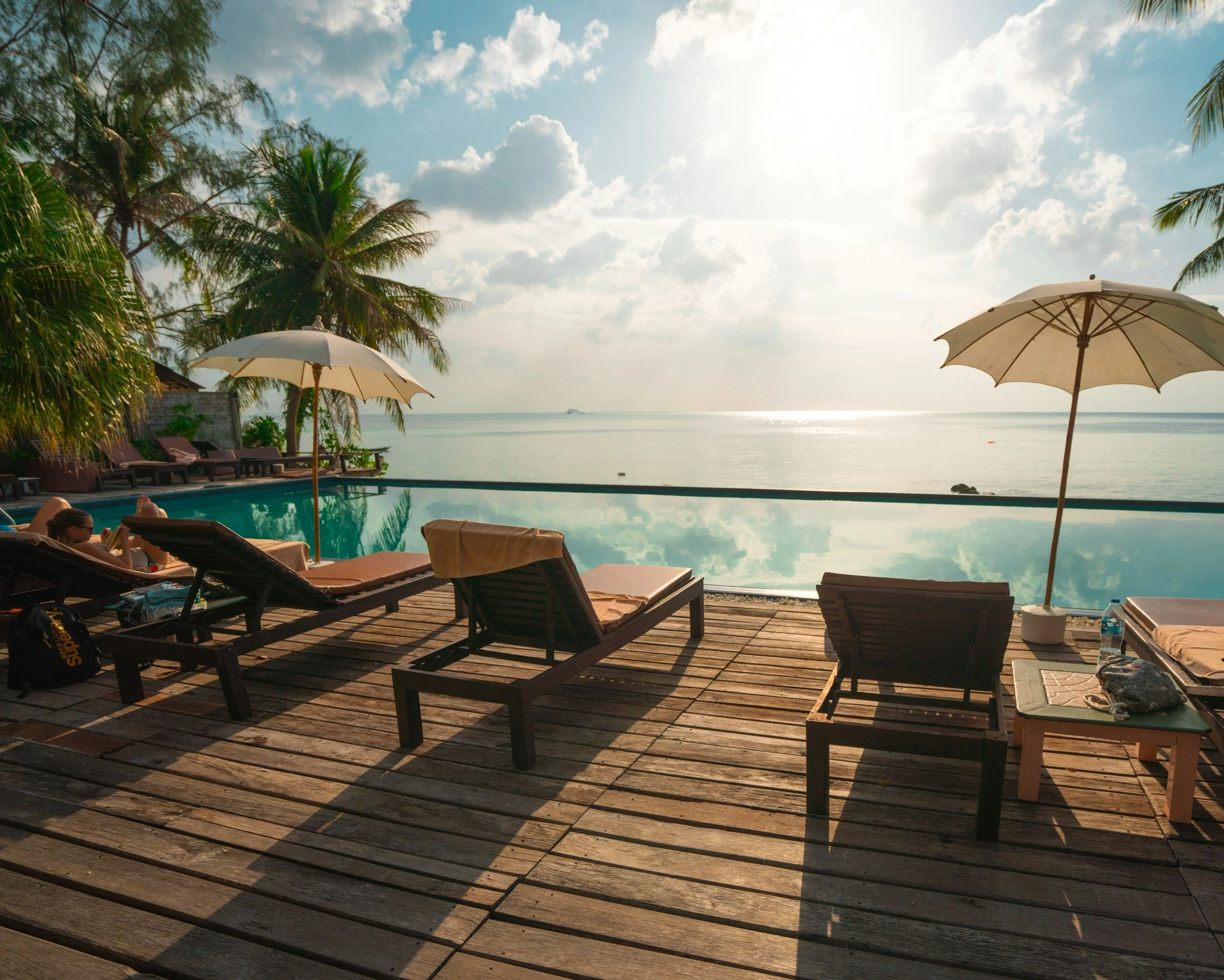 Sunlight shining over a poolside area with lounge chairs, umbrellas, and palm trees overlooking the ocean.