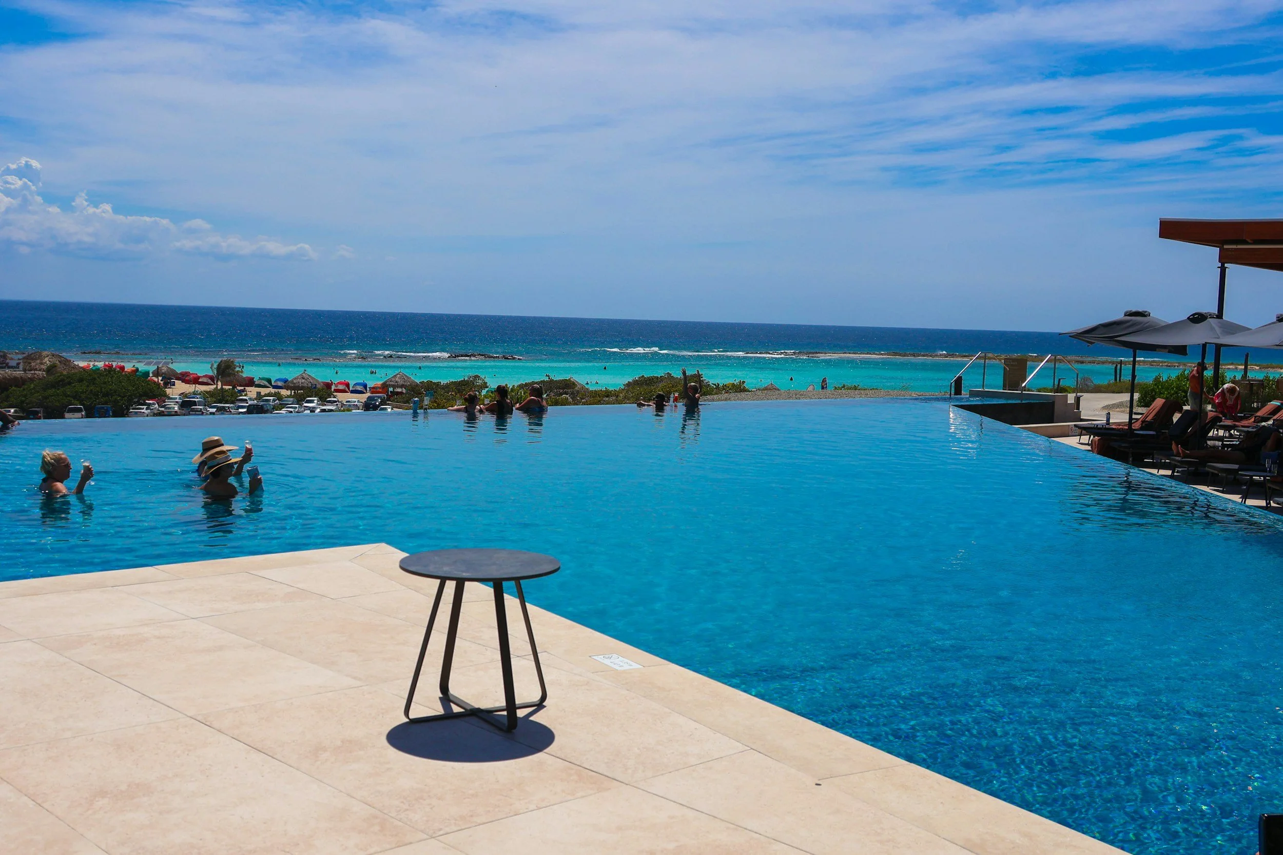 People swimming and relaxing in an infinity pool overlooking the ocean with clear skies and beach umbrellas.