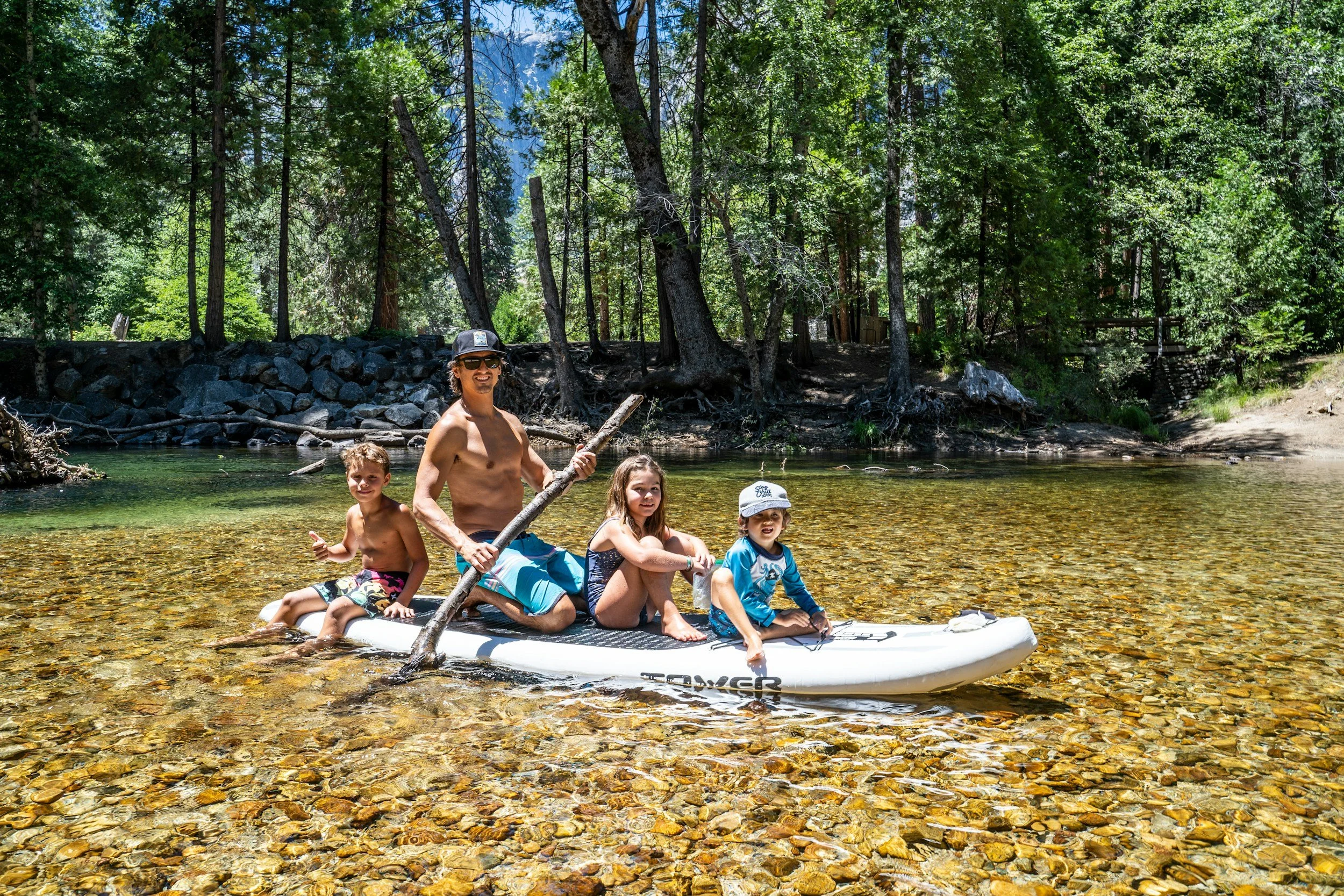 Family of four sitting on a paddleboard in a shallow river surrounded by dense green trees in a forest.