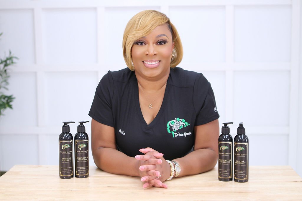 A woman with blonde hair sitting at a table with four black bottles of hair care products in front of her, smiling, against a white background.