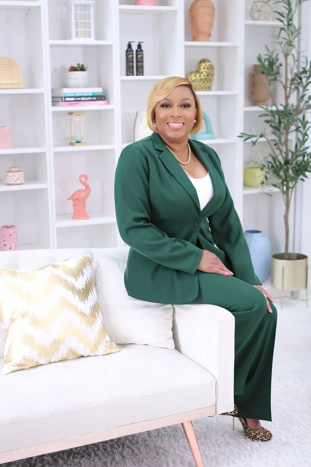 A woman sits on a white sofa in a contemporary interior, wearing a green suit and leopard print shoes, with a smile on her face. Behind her are white shelves with decor items like books, vases, and sculptures, and a potted plant.