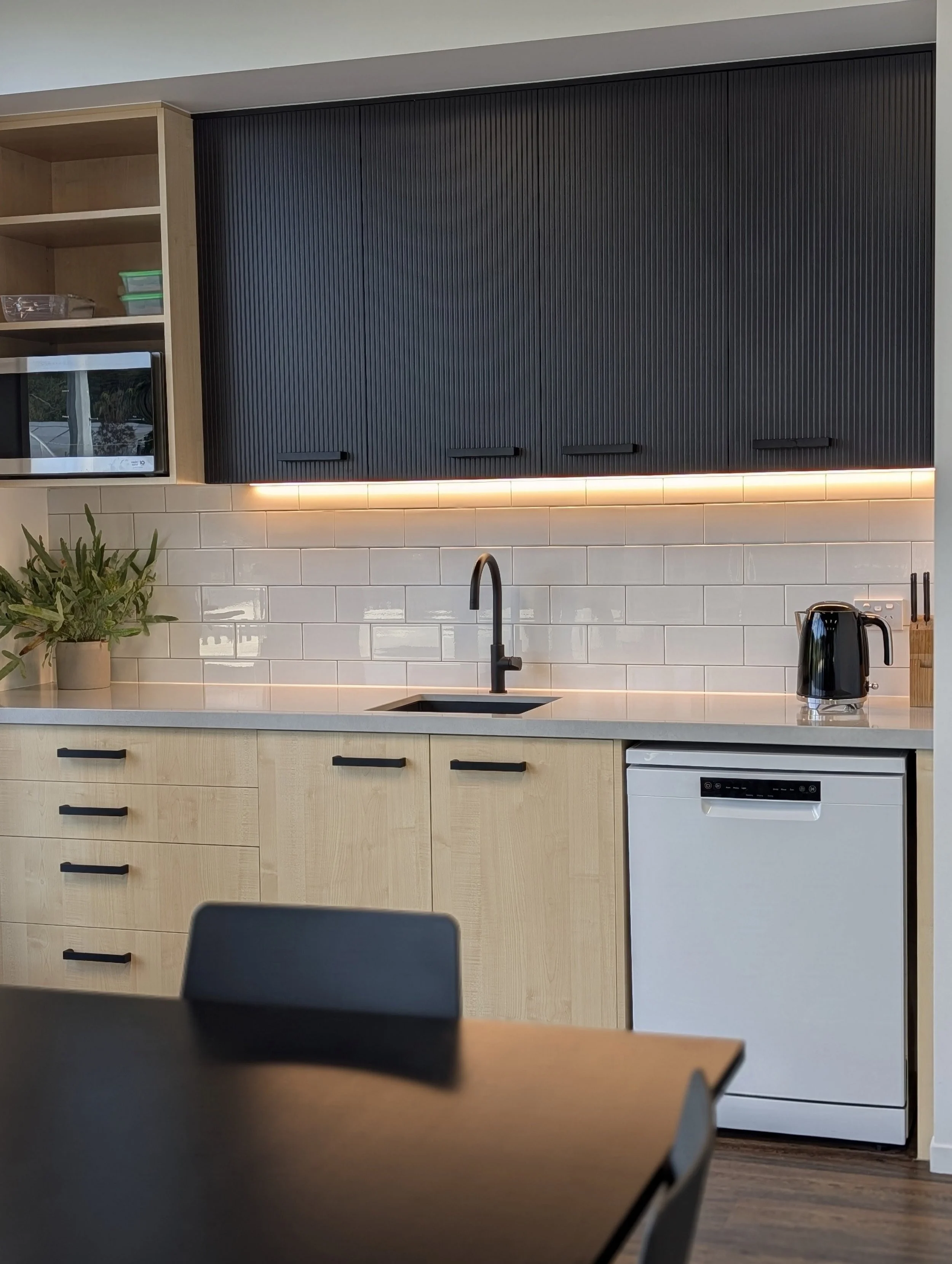 Modern office kitchen with black upper cabinets, white subway tile backsplash, beige lower cabinets, grey countertop with a black sink, a potted plant, microwave and electric kettle.