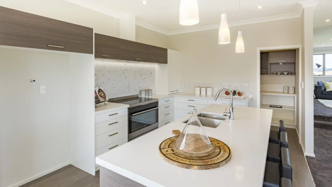 Modern kitchen with white countertops, gray cabinets, and a backsplash with hexagonal tiles, featuring a built-in oven, a sink, pendant lighting, and a view into a living area.