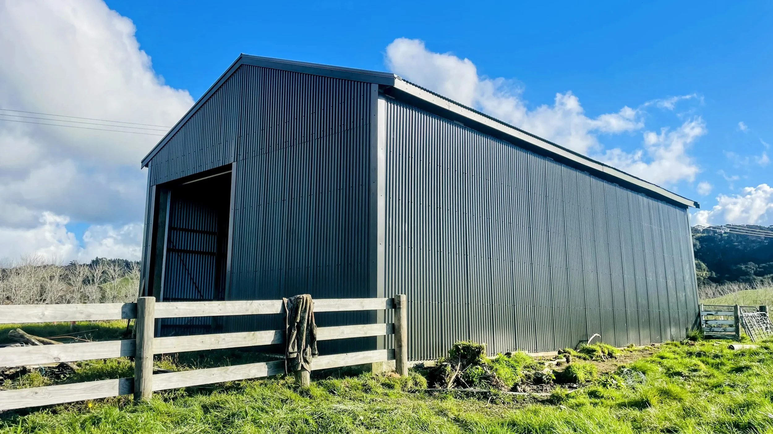Large blue metal barn with open door, surrounded by a wooden fence and green grass, under a partly cloudy sky.
