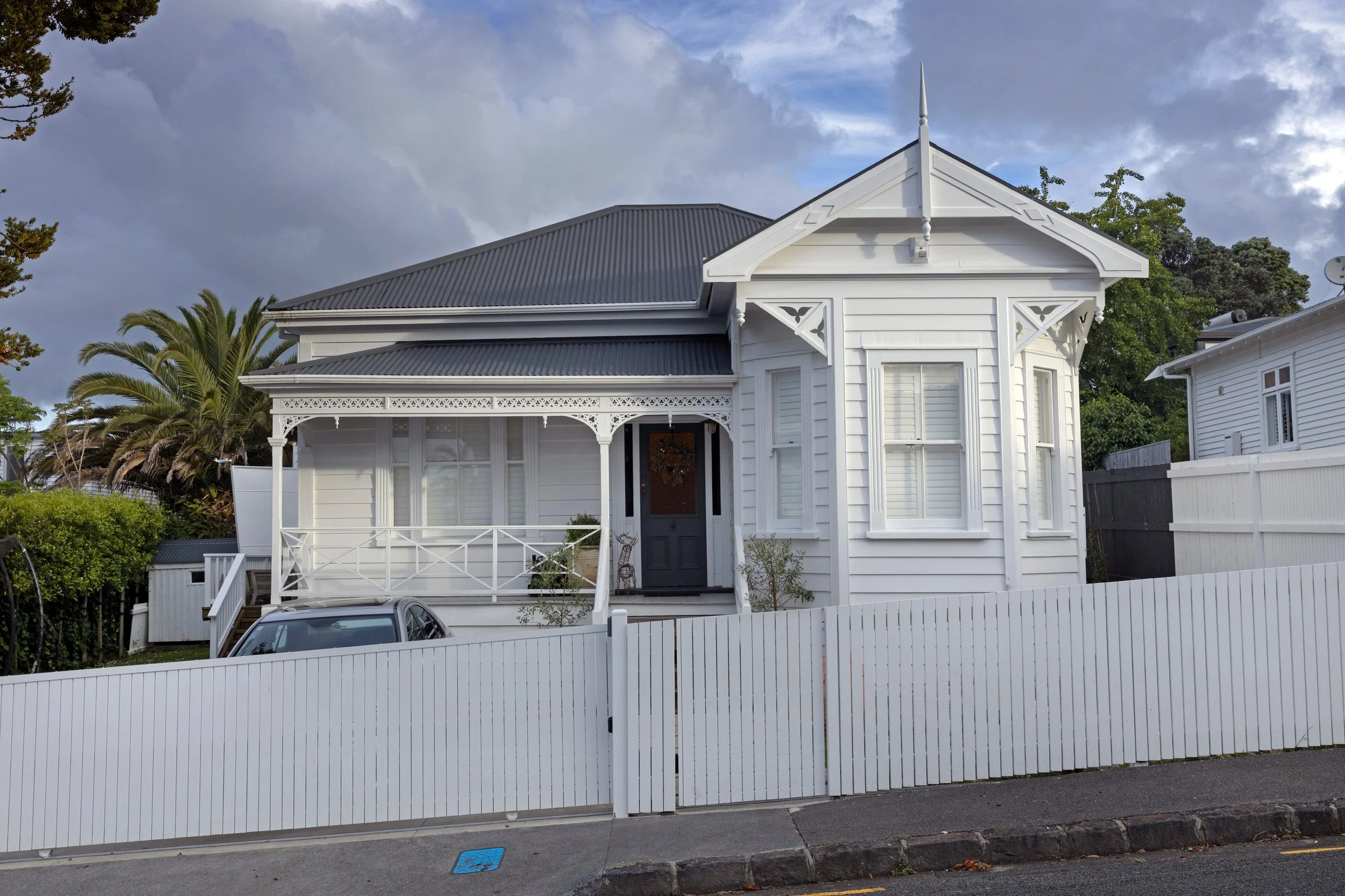 White Victorian-style house with decorative trim, a gray metal roof, and a front porch. A white picket fence surrounds the property, with trees and cloudy sky in the background.