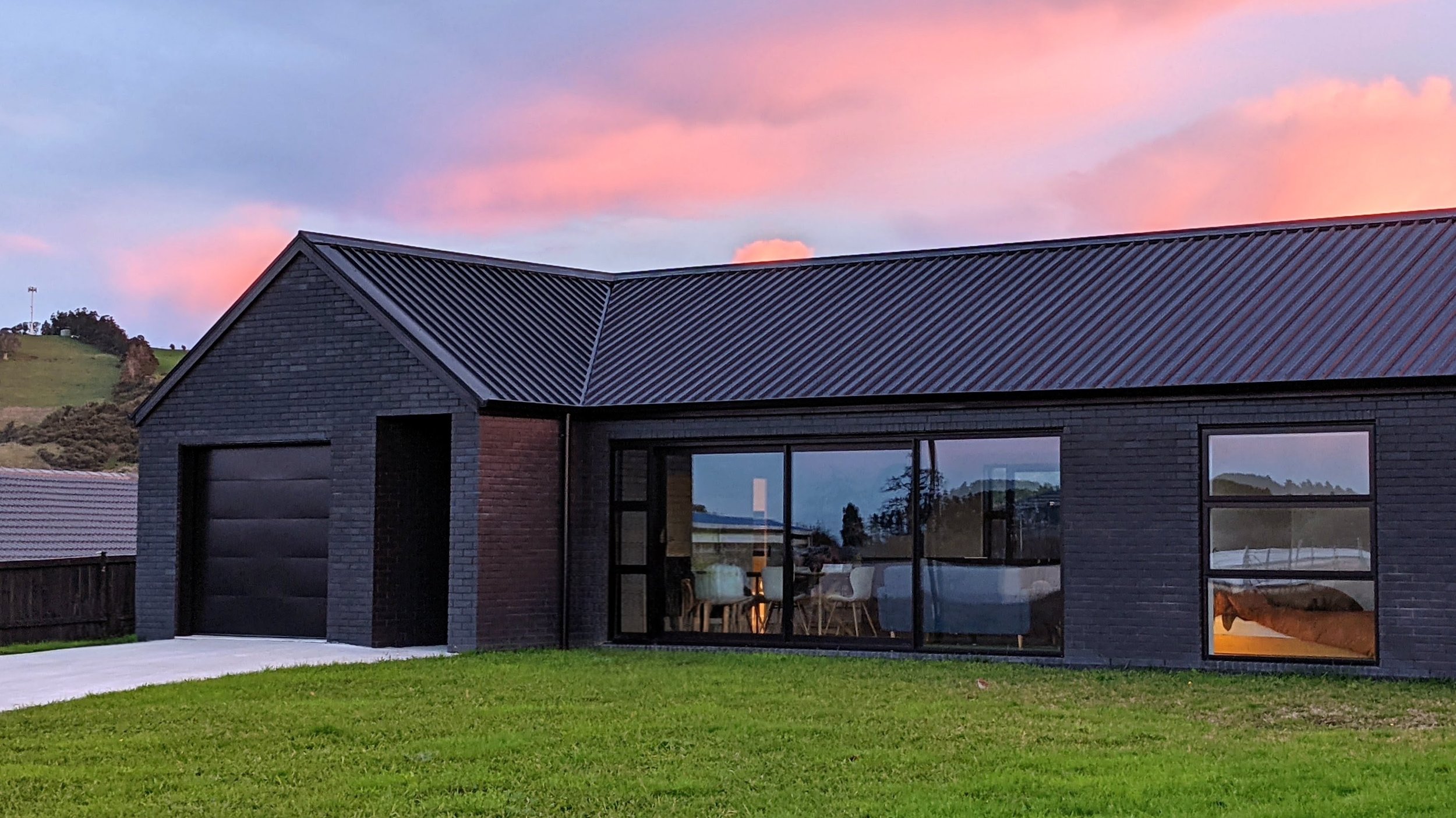 Modern house with black brick exterior, large glass windows, and a metal roof, set against a sunset sky with pink and blue clouds.