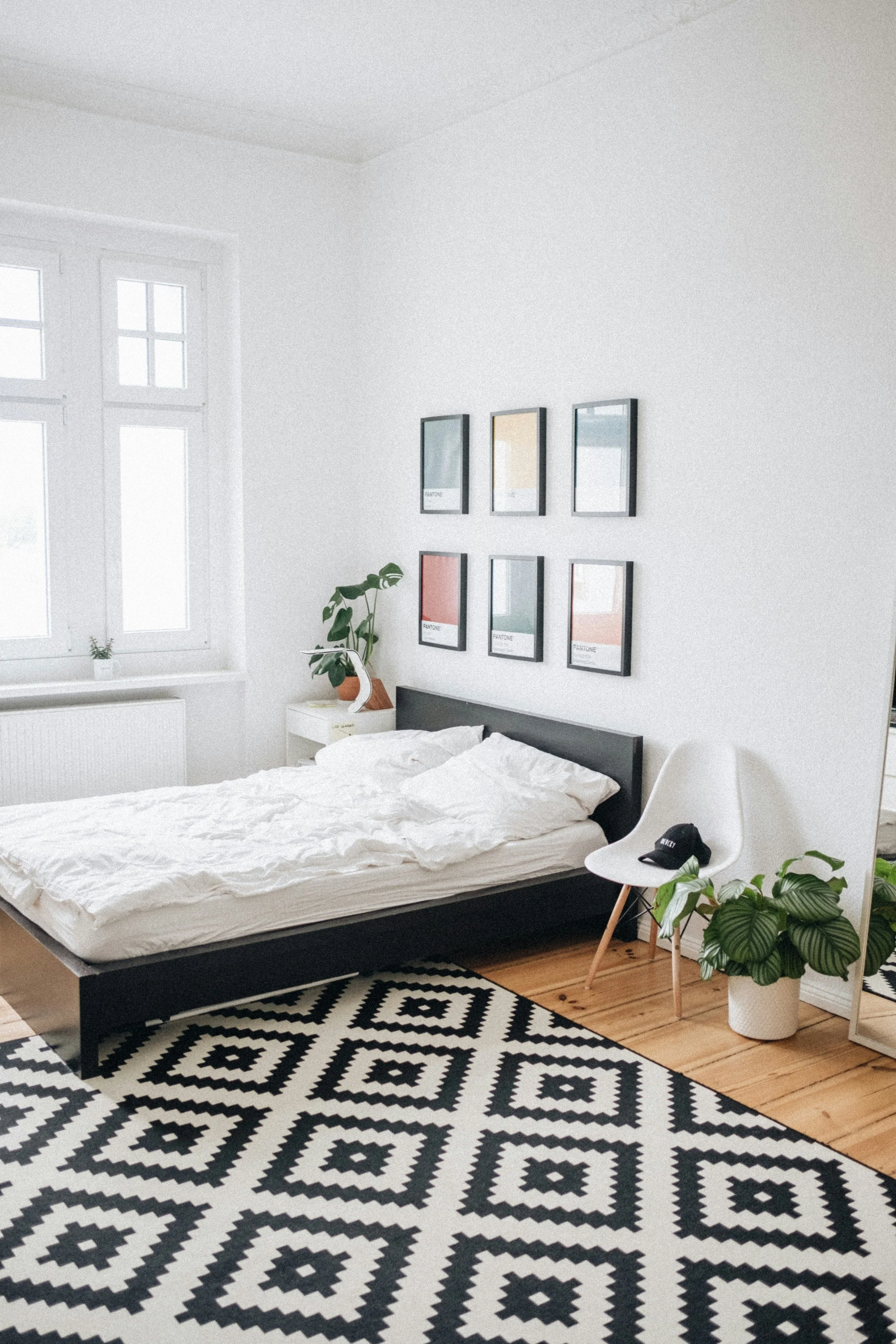 A modern bedroom with a black bed frame, white bedding, and patterned black and white rug. The wall above the bed displays six framed Pantone color swatches. There is a white bedside table with a potted plant. A white chair with a black cap on it and a large potted plant are nearby. The room has white walls, a wooden floor, and a large window with a view outside.