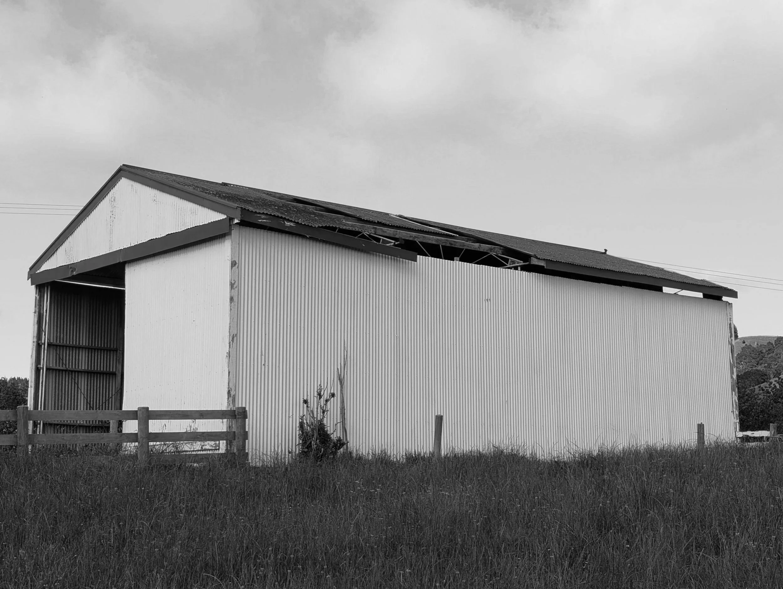 A black-and-white photo of a large warehouse or barn with a sloped metal roof, partially open sides, and a fence in front. The sky is cloudy, and there is grass in the foreground.