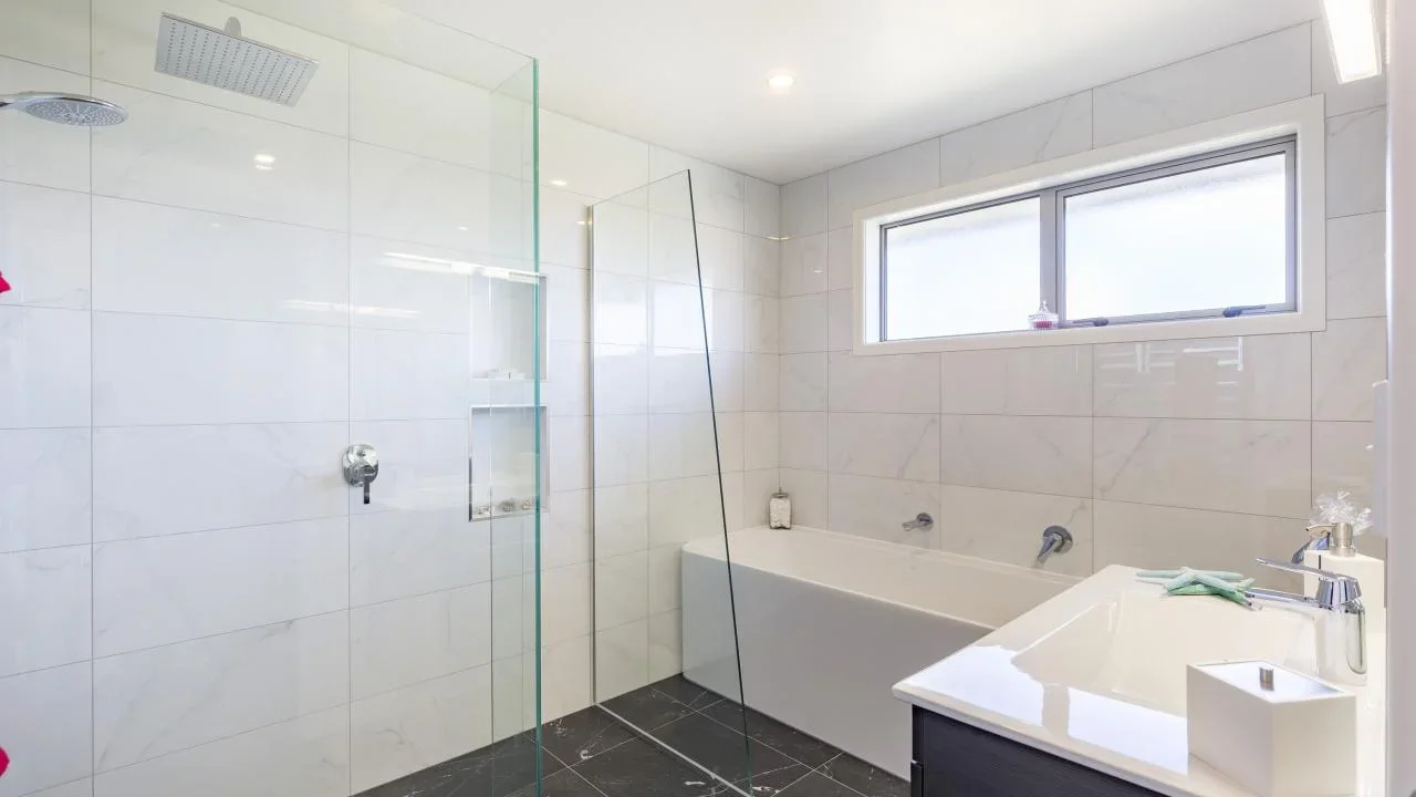 Modern bathroom with white tiled walls, black tiled floor, bathtub, and a walk-in shower area separated by a glass panel, natural light from a window.