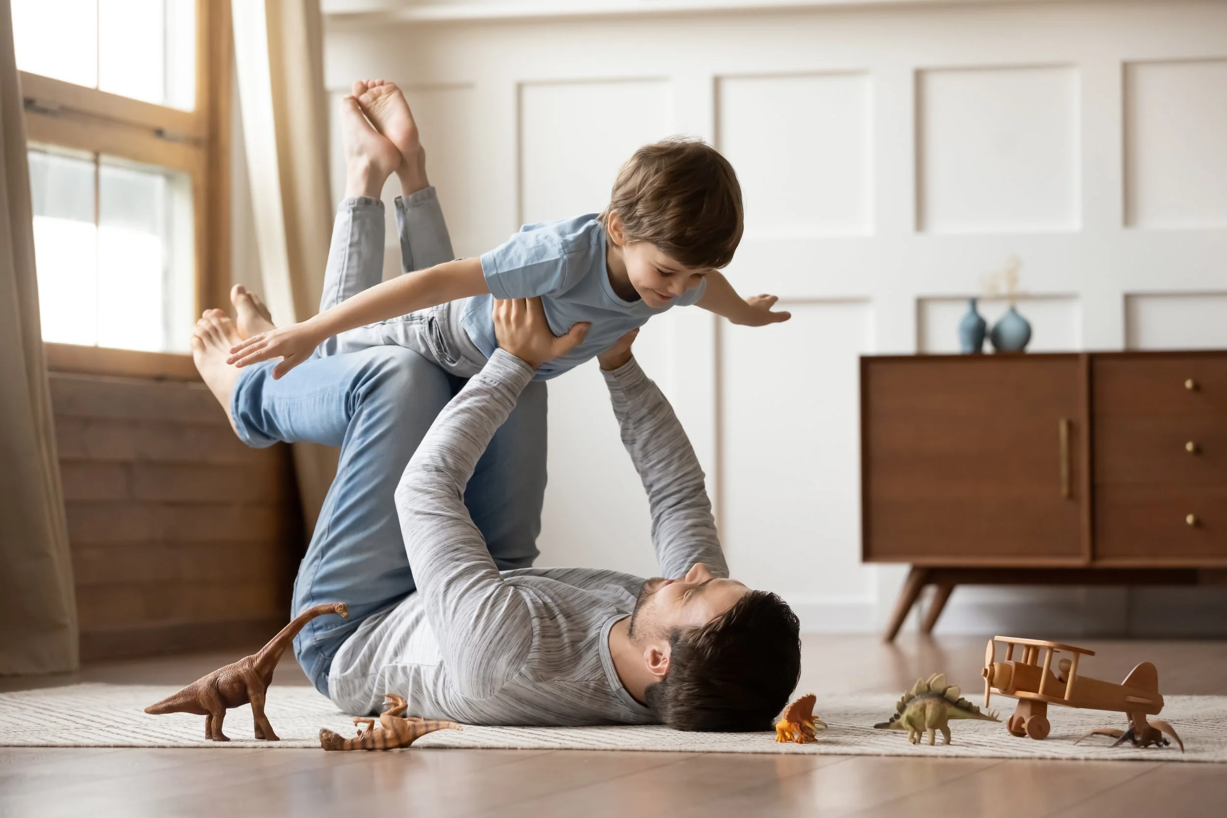 A man on the floor holding a boy in the air while they play with toy dinosaurs.