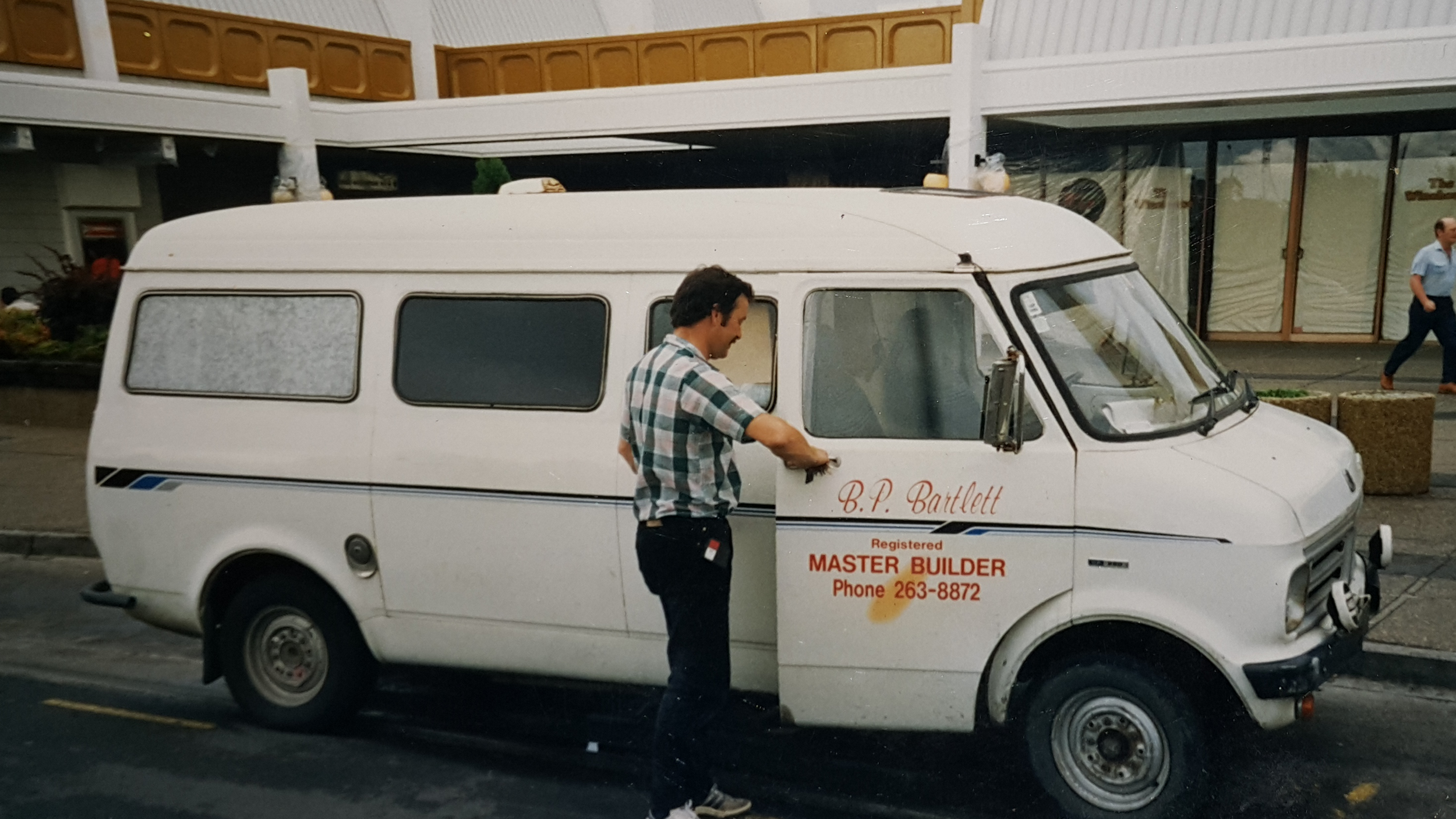 Brian in a plaid shirt is standing next to his white work van with sign writing on the side that reads 'B. P. Bartlett, Master Builder, Phone 263-8872.'