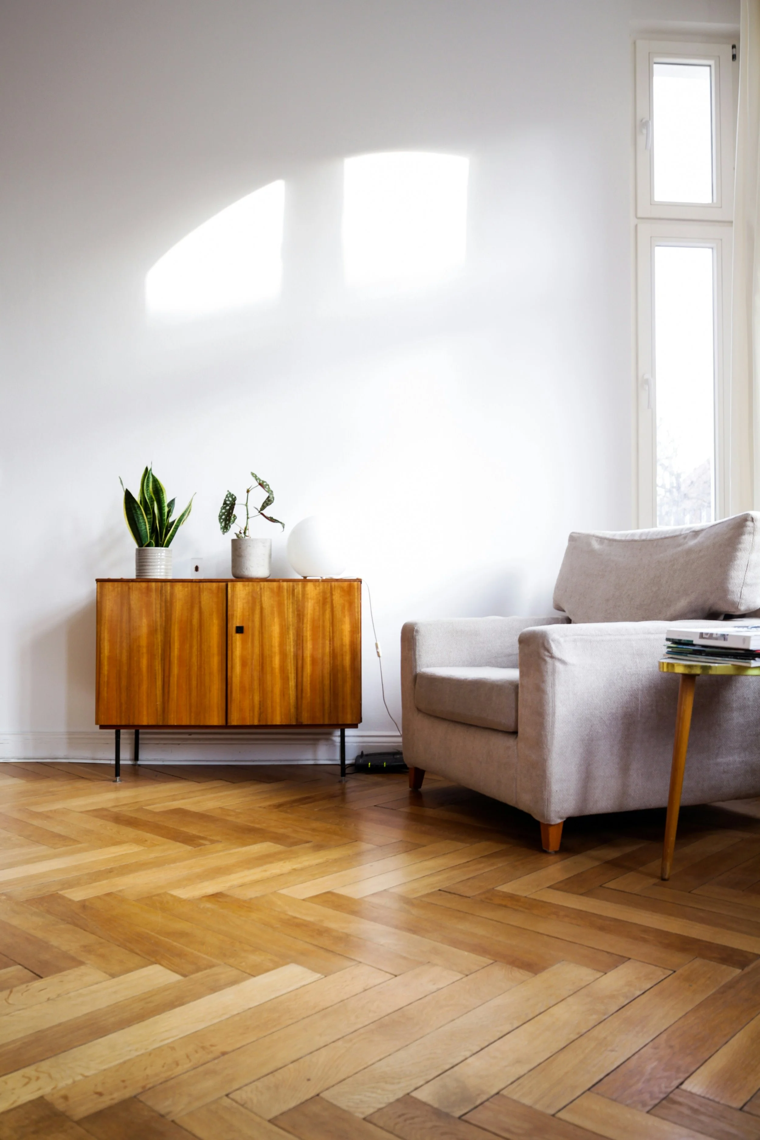 A cozy living room with a light-colored sofa, a small wooden cabinet with black metal legs, and two potted plants on top. Natural sunlight streams through a tall window, casting shadows on a white wall behind.
