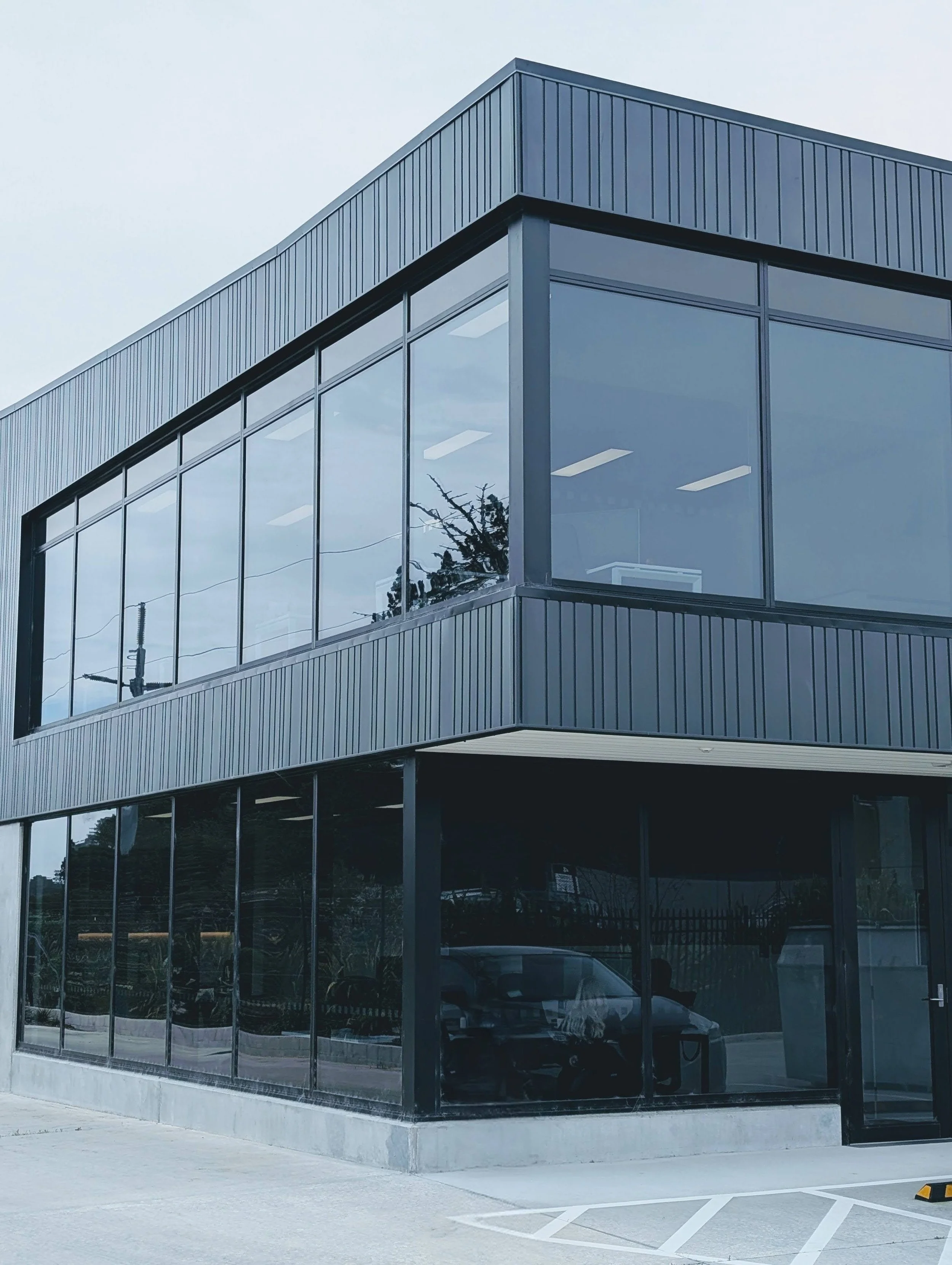 Modern glass-front building with reflecting windows, parked cars, and nearby trees, under a clear sky.