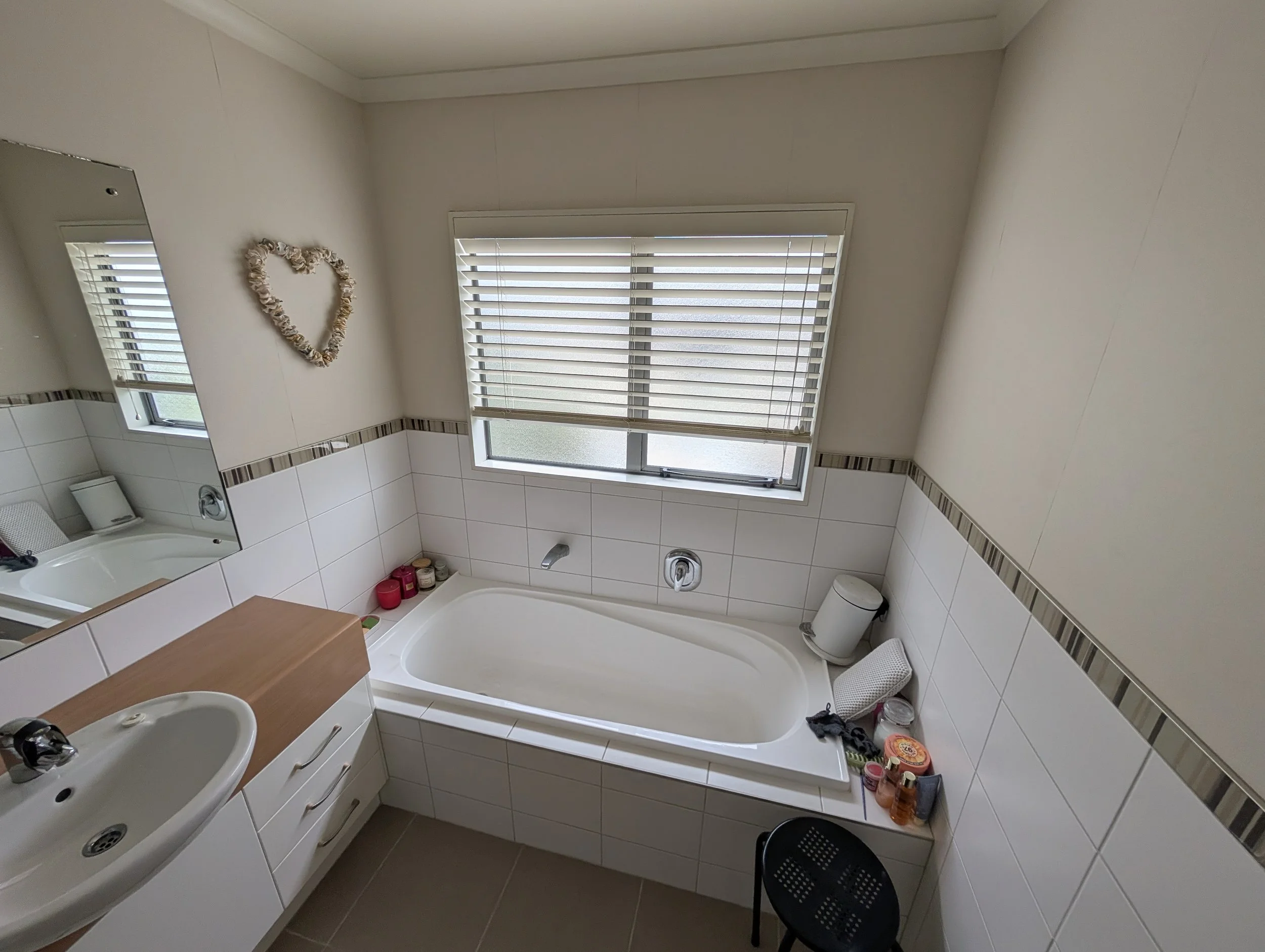A clean bathroom with white tiles, a bathtub, a large window with blinds, a mirror with a heart-shaped decoration, a sink, and various toiletries on the counter and shelves.