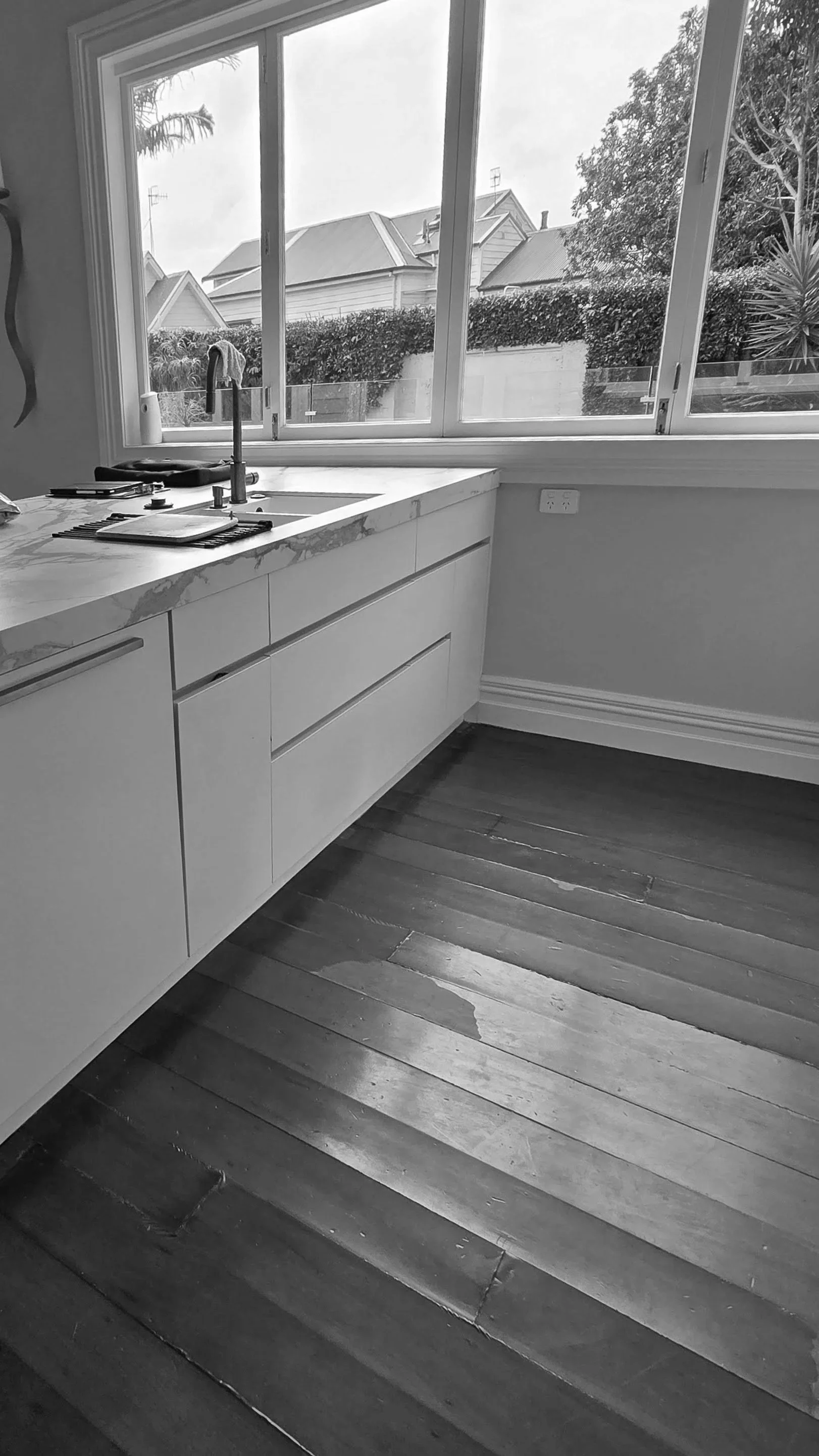 A black and white photo of a kitchen with a large window showing houses and trees outside. There is a white cabinet and a marble countertop with a sink and some items on it, along with wooden floorboards.